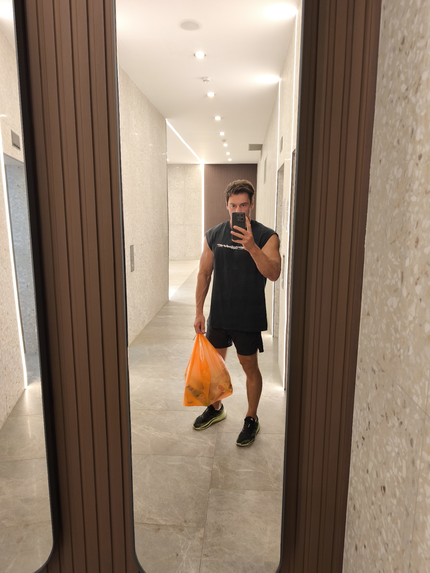 A man in a black sleeveless shirt and black shorts is taking a selfie in a hallway. He is holding an orange plastic bag in his left hand and a smartphone in his right hand. The hallway has white marble floors, and the walls are decorated with beige and white speckled tiles. There are vertical brown panels on the left and right sides of the hallway, which appear to be part of elevator doors or decorative elements. The lighting in the hallway is provided by ceiling lights and LED strips embedded in the walls. The man is reflected in a large mirror on the left side of the frame.