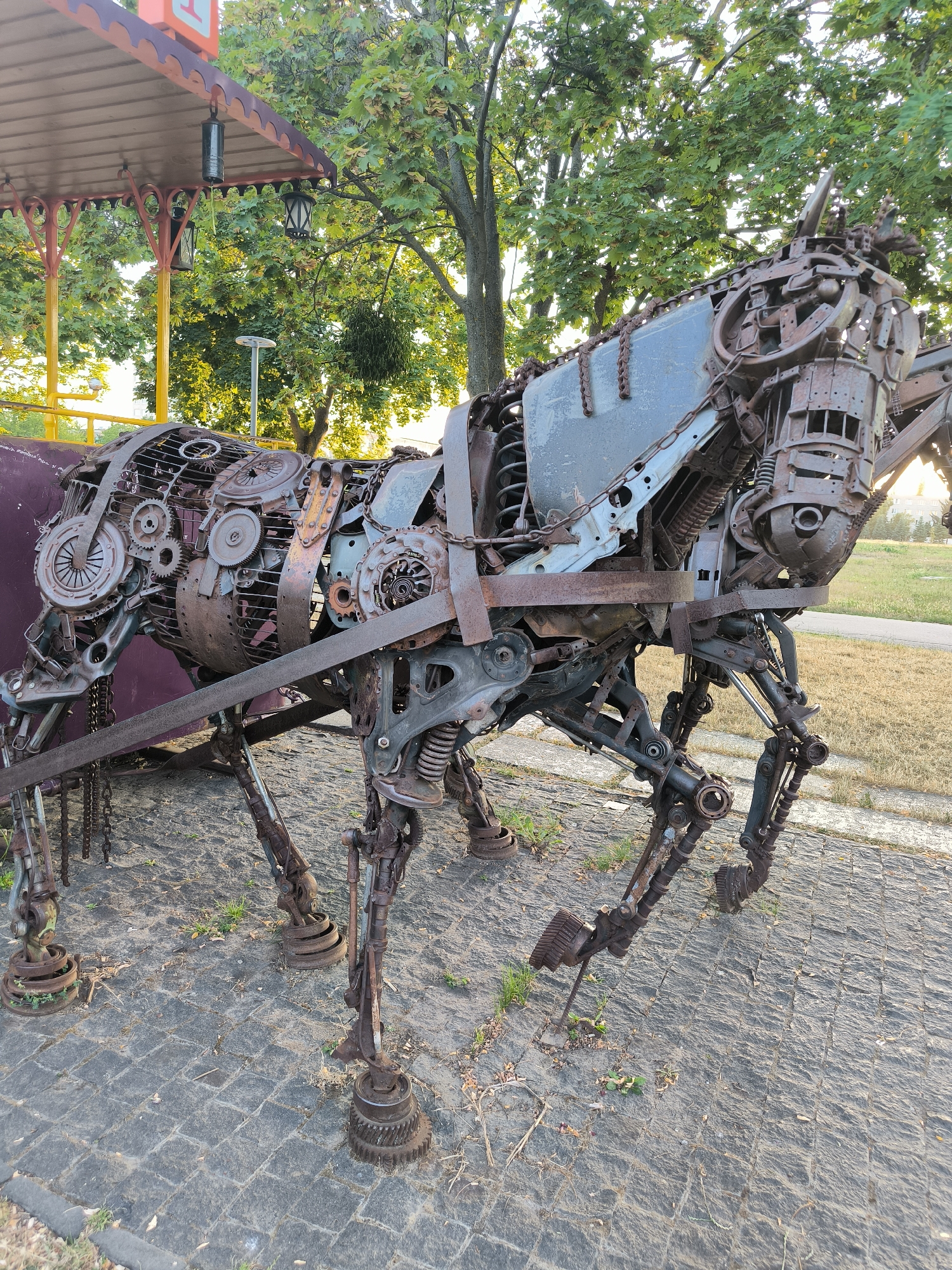 The image displays a large, intricately constructed metal sculpture of a horse, assembled from various salvaged mechanical parts, including gears, chains, springs, and car components. The sculpture is positioned outdoors on a paved area, possibly a walkway or plaza, in Chișinău, Moldova. The horse sculpture is the primary subject, occupying the center of the frame. It is depicted in a standing pose, with its legs positioned as if in motion. The sculpture's body is a complex arrangement of rusted metal, showcasing a steampunk aesthetic. In the background, there are trees with green foliage, suggesting a park or outdoor public space. A portion of an overhead structure, possibly a gazebo or awning with decorative elements and hanging lanterns, is visible to the left. The lighting suggests it is daytime, with sunlight casting shadows. No people are present in the image. The environment appears to be well-maintained, with the paved ground showing some scattered leaves and grass.
