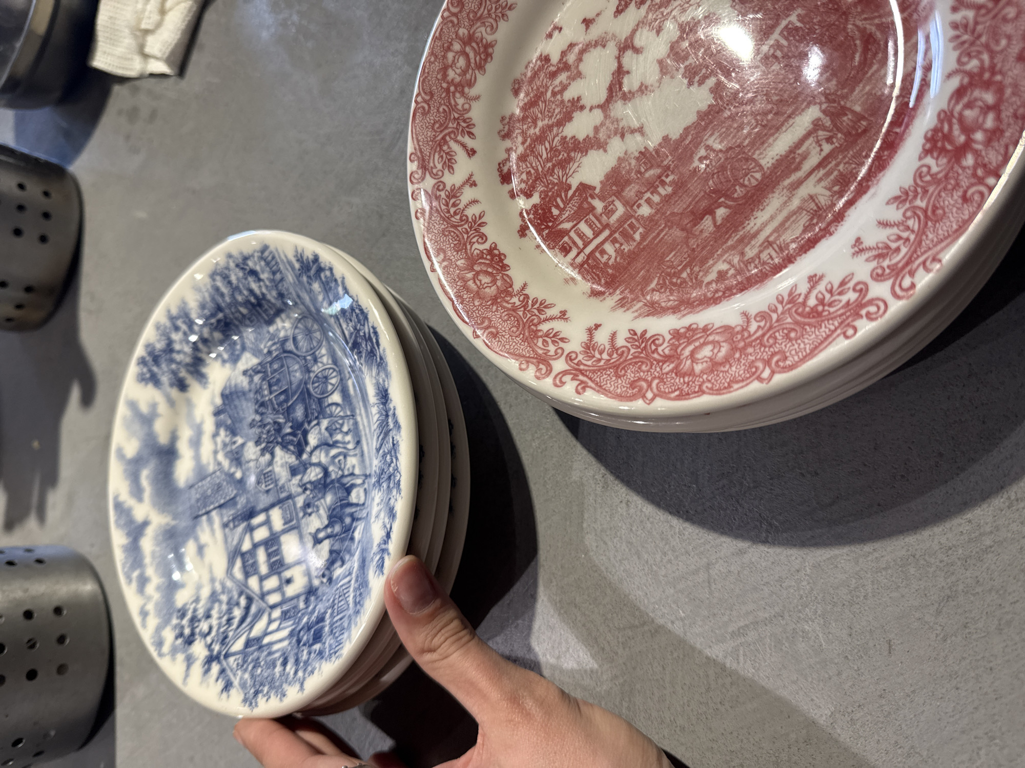 The image displays an indoor scene, likely a kitchen or dining area, featuring two stacks of decorative ceramic plates on a textured grey surface. A human hand, possibly belonging to a woman given its slender appearance and a ring on the index finger, is holding a stack of smaller plates in the lower left portion of the frame.
These smaller plates are adorned with intricate blue and white patterns, depicting a detailed pastoral or historical scene that includes a building and a horse-drawn carriage. To the upper right, a larger stack of plates showcases similar elaborate patterns, but in red and white. These red-patterned plates also feature landscapes with buildings and trees, characteristic of a toile design. Both sets of plates have a glossy finish, which reflects ambient indoor lighting, creating highlights.
In the background, to the far left, a metallic object with perforations, possibly a utensil holder, is partially visible. Further back, a white cloth or towel and part of a metallic pot can be indistinctly seen. The activity captured is the handling or arrangement of these plates. The image conveys a sense of domesticity, highlighting classic or vintage dinnerware. There is no visible text in the image. While provided with the context of Izhevsk, Russia, there are no specific visual cues within the image itself that identify this location.