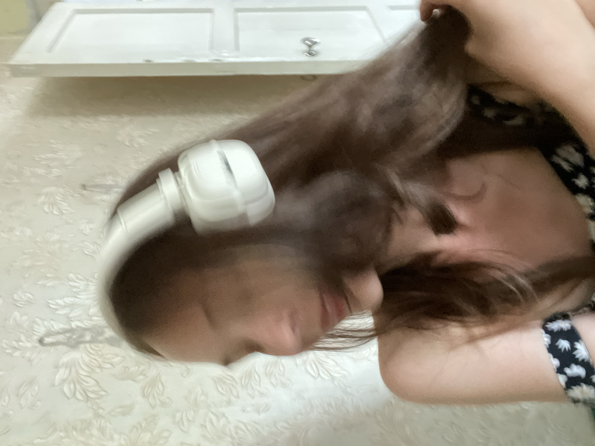 The image depicts a medium shot of a young woman with long, brown hair wearing white headphones. She is positioned in what appears to be a domestic indoor setting. The background features textured wallpaper with a floral pattern and a white door. The lighting is soft, and the woman's face is slightly blurred due to motion or focus. She is looking down and to her left, with her right hand near her head. Her left shoulder and arm are visible, and she is wearing a dark top with a white floral pattern. The overall impression is a candid, casual moment.