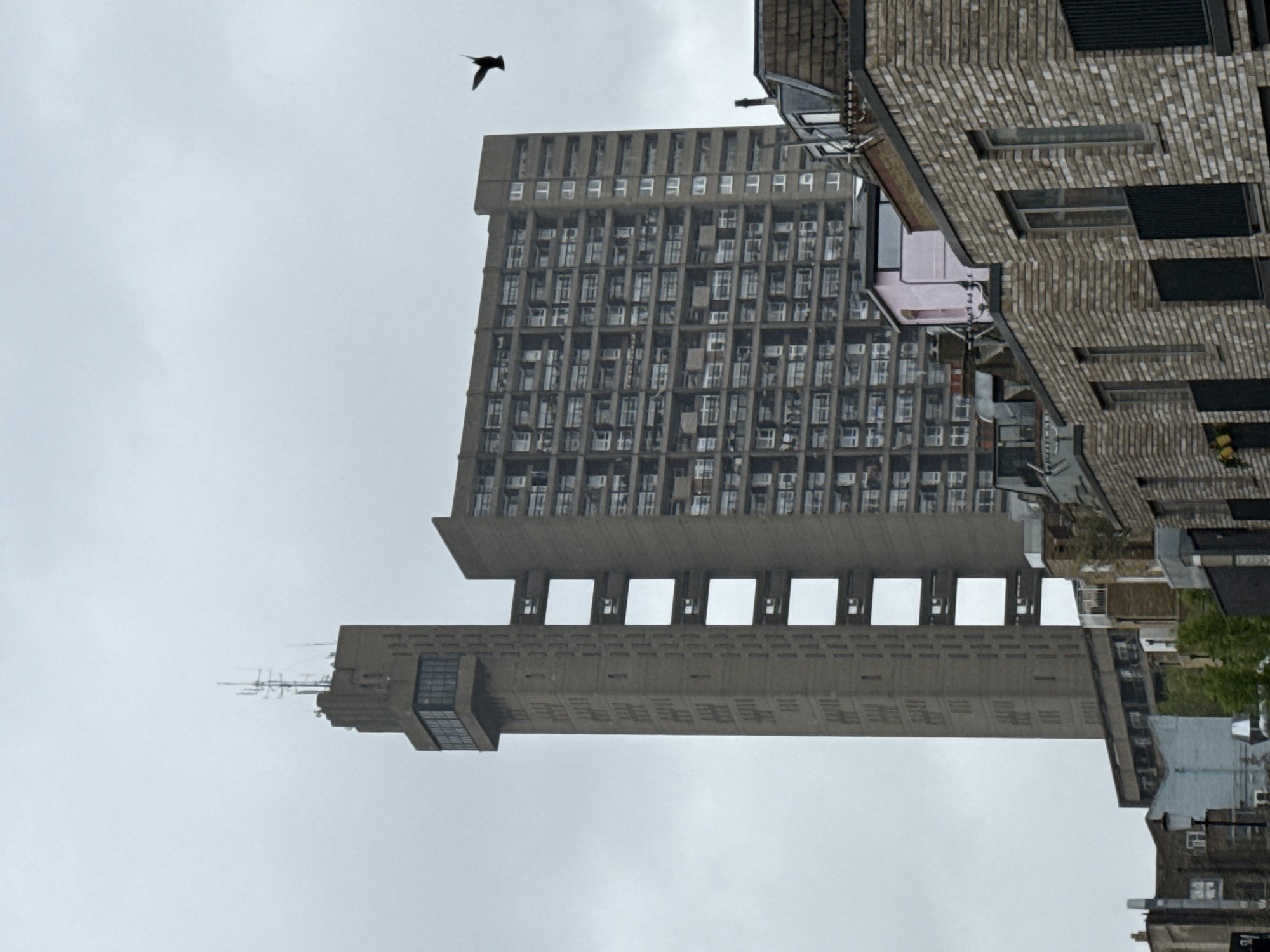 The image depicts the Barbican Estate in London, United Kingdom. The Brutalist architecture is prominent, showcasing the complex's concrete towers and residential blocks. A bird is flying above the buildings. The sky is overcast. In the foreground, additional buildings, including a brick structure, are visible.