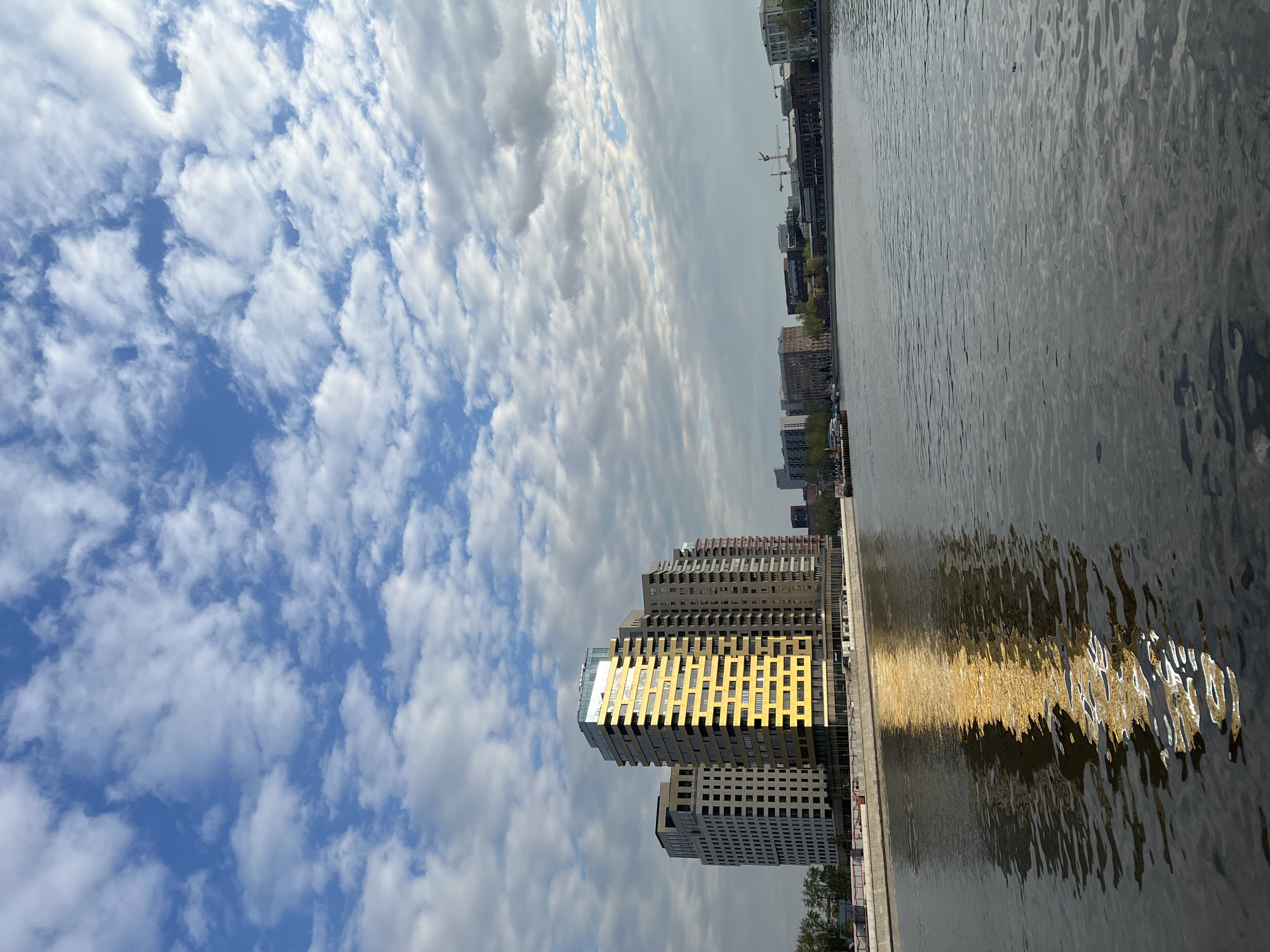 The image displays an urban waterscape, likely in Moscow, Russia, under a bright, partially cloudy sky. A wide body of water, presumably a river, occupies the right and lower-right sections of the frame, its surface rippled and reflecting sunlight.
On the left side, a cluster of modern high-rise buildings stands prominently. One building features distinctive golden or yellow-colored facade sections that brightly reflect the sun. This golden glow is mirrored vividly in the water, creating a shimmering, distorted reflection. Other buildings in the cluster exhibit contemporary designs with numerous windows and various tones of grey and beige.
The upper-left portion of the image is filled with a clear blue sky dotted with scattered white clouds, indicative of a sunny day with dynamic weather patterns. Across the river, a distant city skyline is visible, comprising more urban structures and at least one visible construction crane, suggesting ongoing development. Patches of greenery, possibly trees, can be observed along the distant riverbanks.
The time of day appears to be daytime, characterized by strong sunlight. There is no visible text in the image.