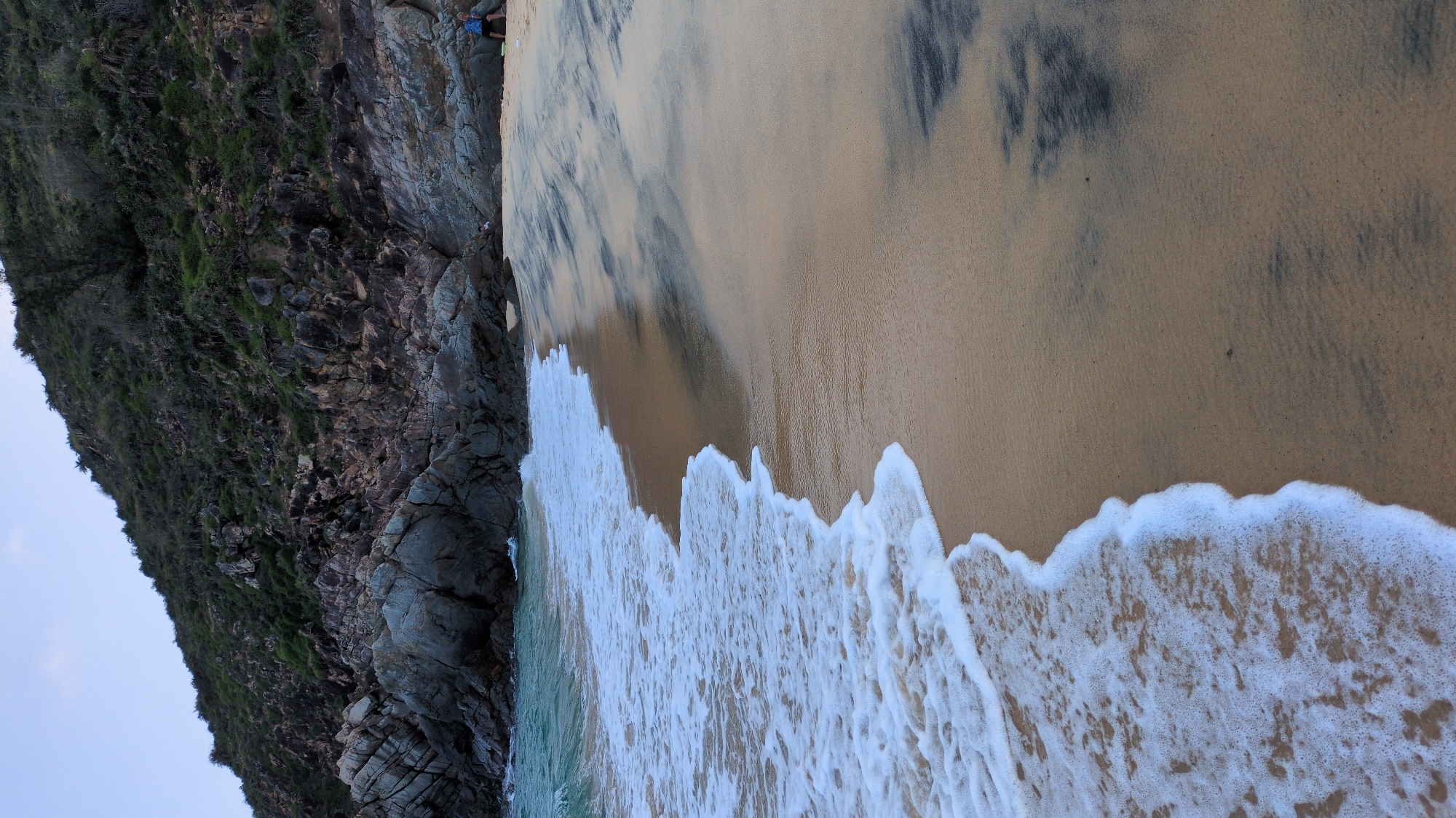 The image displays a coastal scene in East Quy Nhon Ward, Vietnam. A light brown sandy beach occupies the foreground, meeting the ocean water with white foamy waves breaking and receding. Irregular dark grey and black streaks are visible across the wet sand. The ocean, characterized by turquoise-blue water, extends into the midground. The background features a steep, rocky hillside composed of large grey boulders and darker, rugged outcrops, partially covered with sparse green vegetation, predominantly shrubs and small trees. One individual is present on the beach in the far background, positioned against the base of the rocky hill on the right side of the frame, appearing to be seated or crouching while wearing a blue garment.