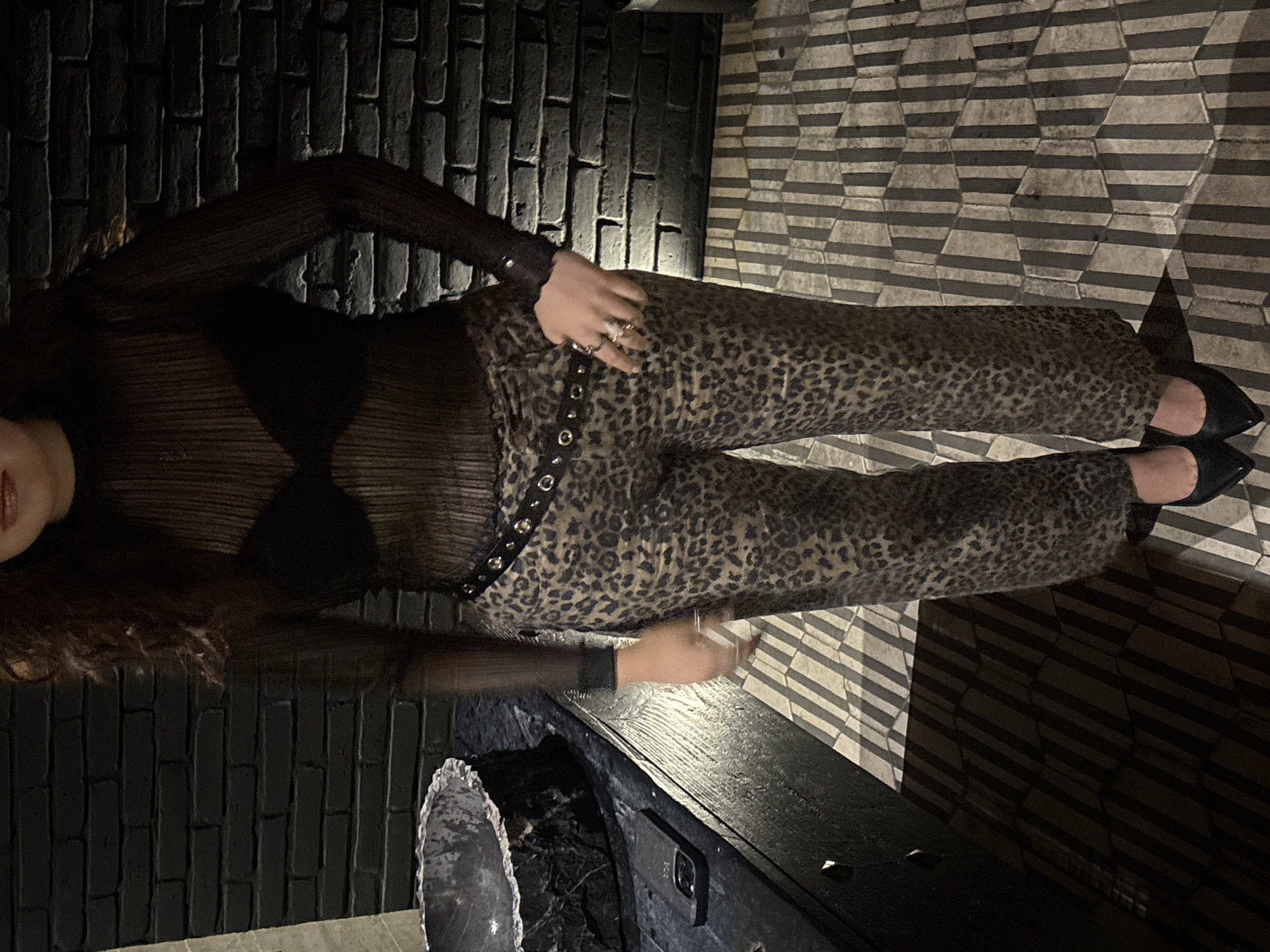 A woman in a sheer black long-sleeved top, a black bra, leopard print pants with a studded belt, and black high heels is positioned in a dimly lit interior, likely a bar or lounge. She is standing next to a dark-colored counter or ledge that has a decorative rock or crystal on top. The background consists of two different patterns; a dark faux brick wall on the left and a geometric patterned floor on the right, suggesting the edge of a tiled area. The image was taken indoors, likely in San Andrés Cholula, Mexico.