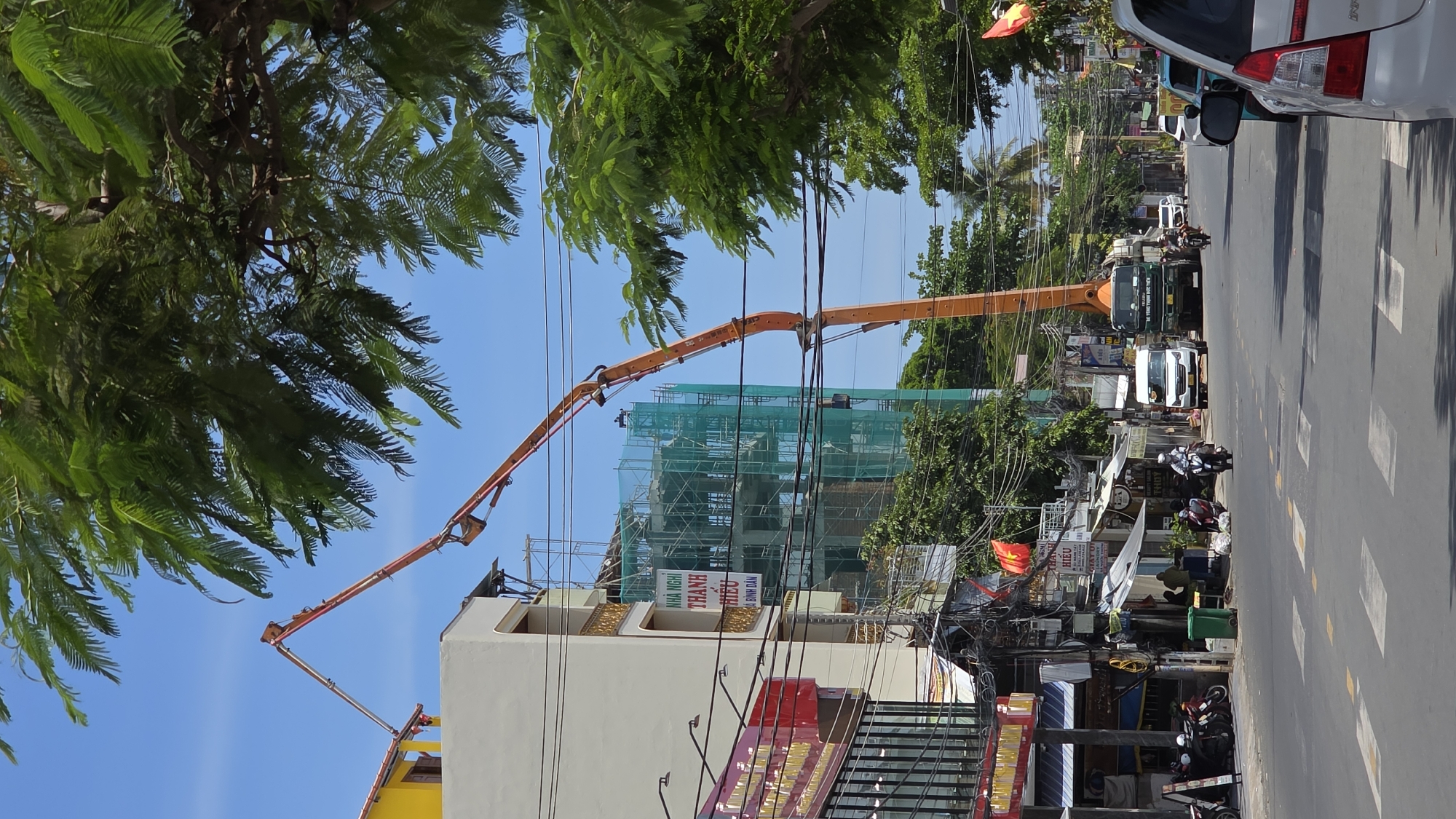 The image shows a street scene in Nha Trang, Vietnam. An orange boom crane is positioned next to a building under construction, covered in scaffolding and green netting. The building is several stories high, partially painted yellow and white. The boom crane's base is a dark green truck parked on the street. In the foreground, power lines run across the sky. The street has a crosswalk, and there are motorbikes and vehicles in the background. A Vietnamese flag is visible near the street. There are businesses on the street. The area appears urban and busy.