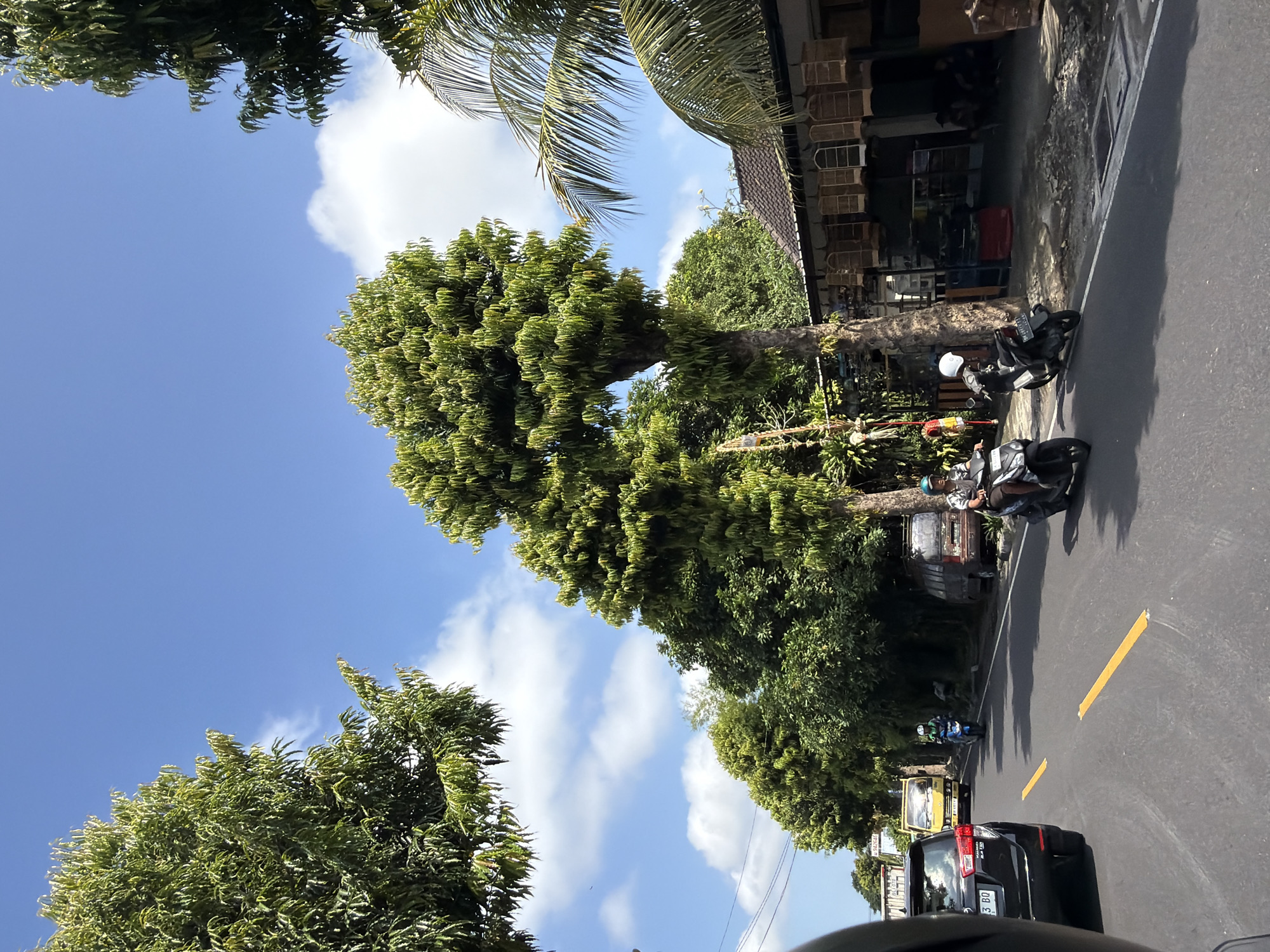 The image depicts a sunny day on a road in Blahbatuh, Indonesia. Two individuals are riding motorbikes on the asphalt road, which has yellow lane markings. A black SUV is in the foreground to the left, and a yellow truck is further ahead on the right side of the road. Lush green trees line the sides of the road, with some buildings visible in the background. One building appears to be a shop selling bird cages. The sky is blue with scattered white clouds.