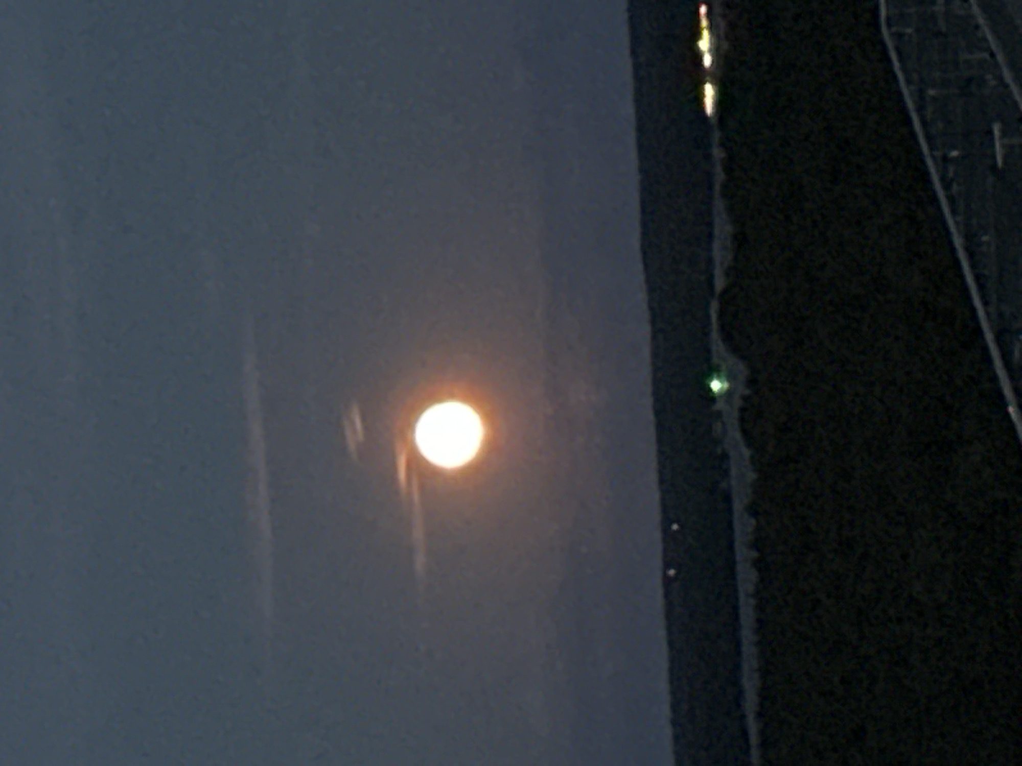 The image shows a full moon in a dark sky above a dark landscape. The moon is bright and appears to be near the horizon. The landscape below is a dark mass of trees or hills, with some lights visible in the distance. There is no indication of the city of Berlin, Germany, or any specific location within it.