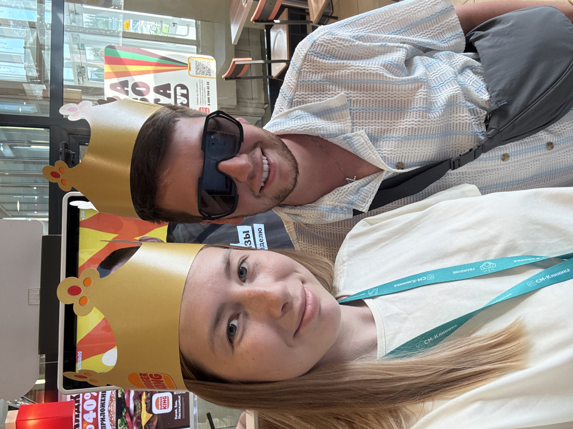 The image depicts a selfie of a man and a woman inside what appears to be a Burger King restaurant. Both are wearing Burger King paper crowns. The man, wearing sunglasses and a patterned shirt, smiles at the camera. The woman, with blonde hair and a lanyard around her neck, also smiles. The lanyard has the text "СМ-Клиника" visible on it.
In the background, a Burger King menu board and promotional materials are visible. A price of "40%" is prominently displayed, alongside text in Russian. Outside, a window shows a glimpse of the city. The phone number “8 800 700 32 32” is visible, along with the name “BURGER KING” and text in Russian. Given the Russian text, "СМ-Клиника" on the lanyard, and the price and menu items, it is highly probable that the location is in the city of Moscow, Russia.