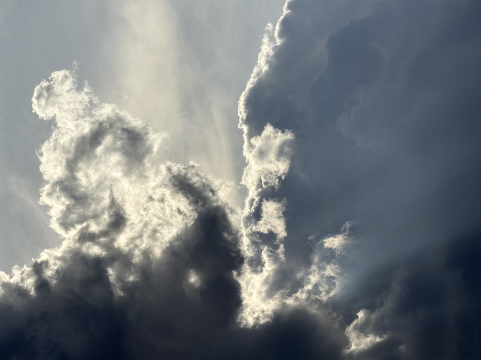 The image is a close-up shot of clouds in the sky. The clouds are a mix of light and dark gray, with some areas appearing white due to sunlight. There are rays of light shining through the clouds. The photograph appears to be taken during the day. The location is not discernible from the image, nor can its location in Krasnoyarsk, Russia be determined.