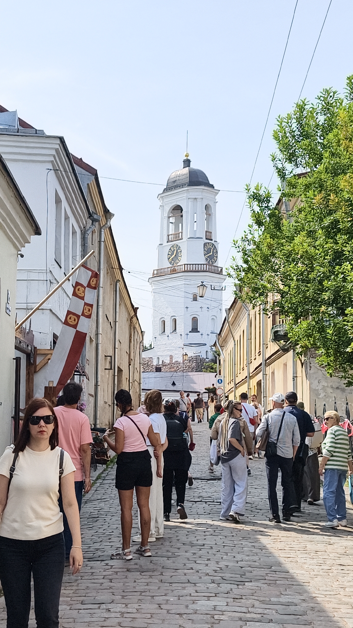 The image depicts a sunny day in Vyborg, Russia, showcasing a cobblestone street lined with historic buildings. In the background, the iconic Clock Tower of Vyborg Castle stands prominently, its white facade adorned with large clock faces. A diverse group of people, dressed in casual summer attire, are walking along the street, some appearing to be tourists. The atmosphere is lively and bustling, suggesting a popular area for exploration and sightseeing. The architecture and setting point to a European city with a rich history.
