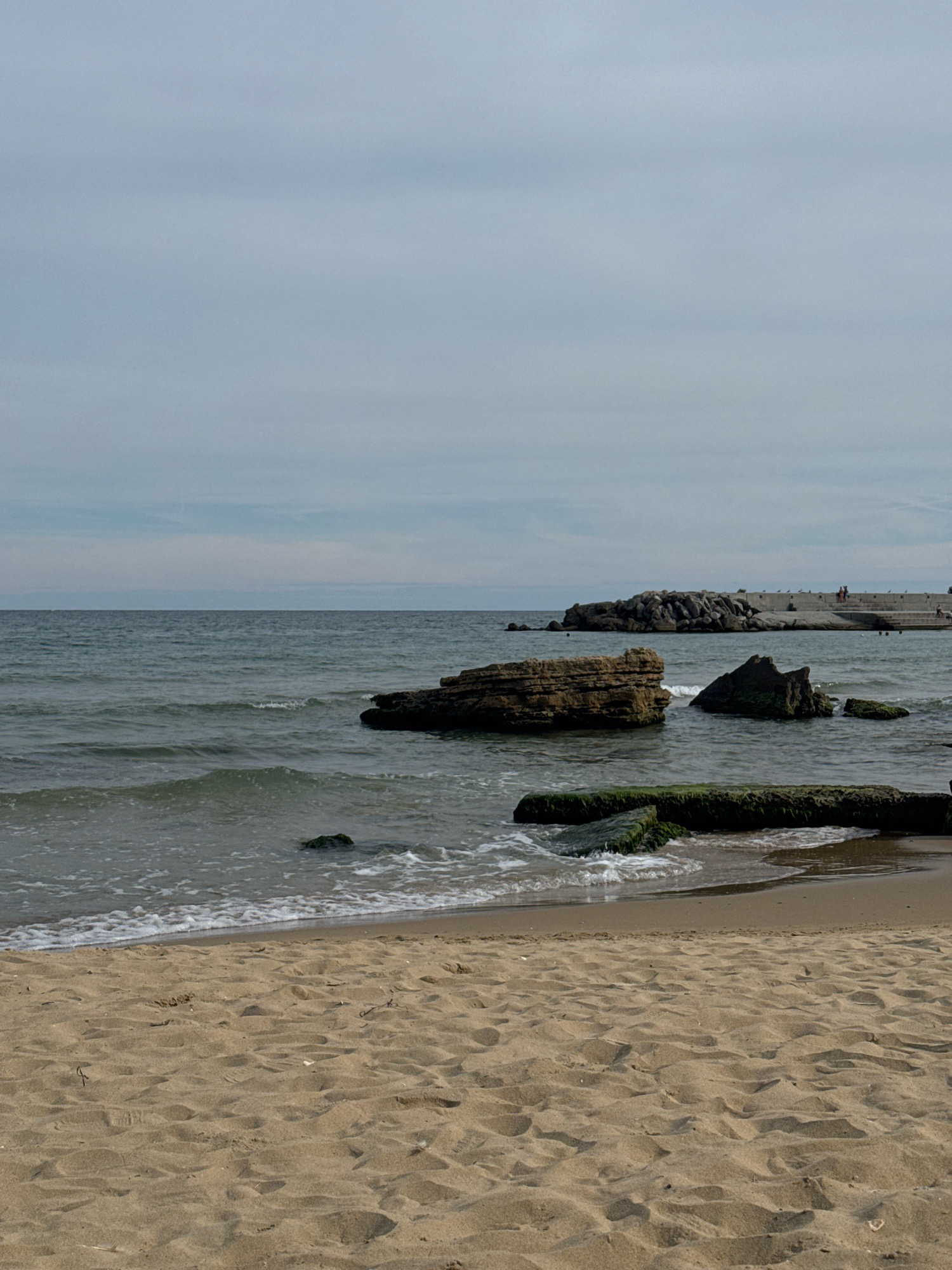 This is a vertical, medium-shot photograph of a beach scene under a cloudy sky. In the foreground, the sandy beach is textured with footprints and scattered debris. The middle ground shows the calm ocean with gentle waves lapping at the shore, creating white foam. Several dark, moss-covered rocks are partially submerged in the water, with one larger, layered rock formation closer to the left. In the background, a concrete breakwater extends into the ocean, with a rocky structure at its base. A few small figures, appearing to be people, are visible on the breakwater, suggesting a public area. The overall environment is that of a coastal landscape.