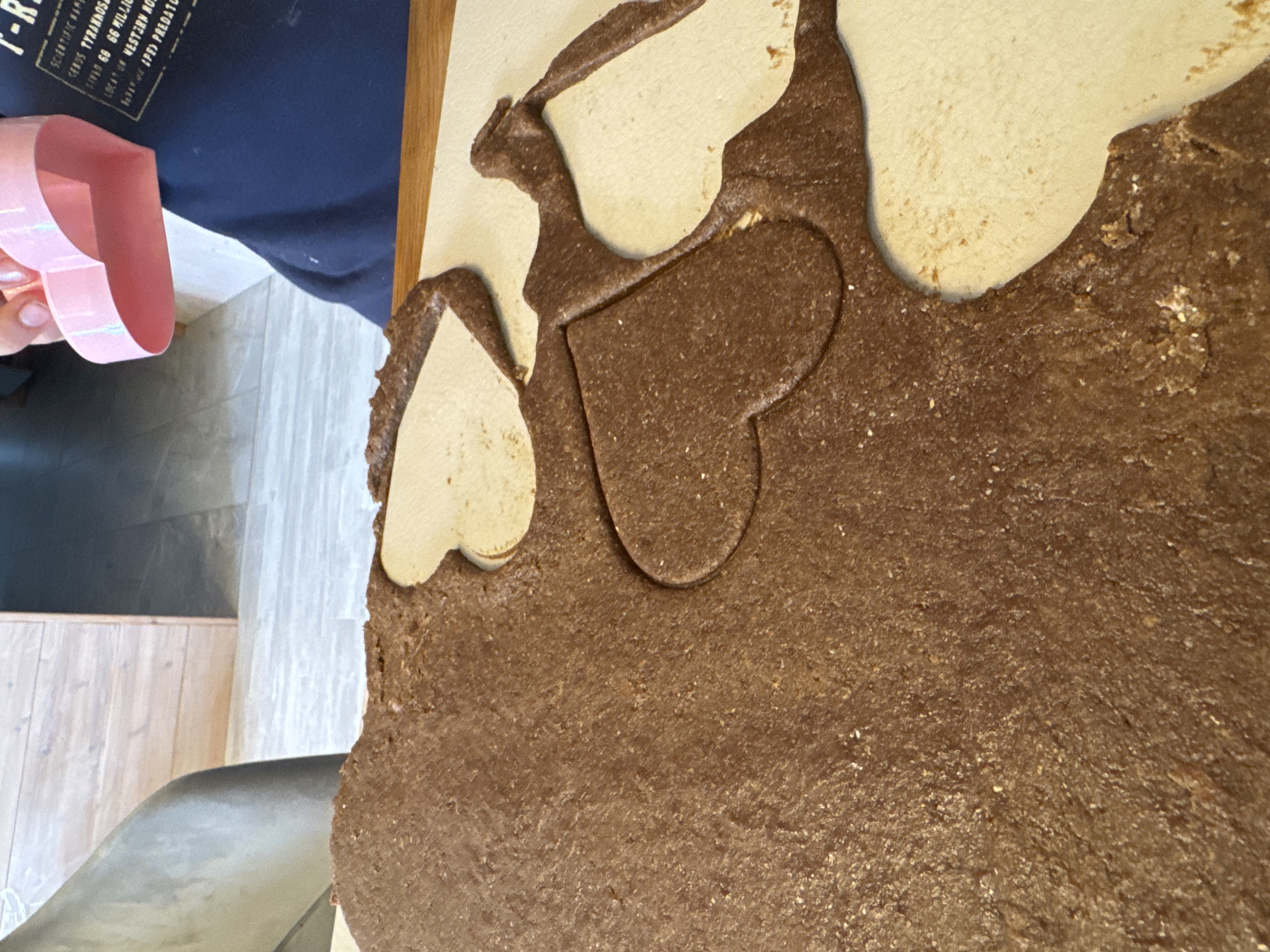 A close-up shot depicts a person using a pink heart-shaped cookie cutter on rolled-out chocolate cookie dough. The dough rests on a wooden surface. Part of the person's navy blue shirt with text on it is visible. In the background is a room with light-colored wooden floors. The location is impossible to determine based on the image.