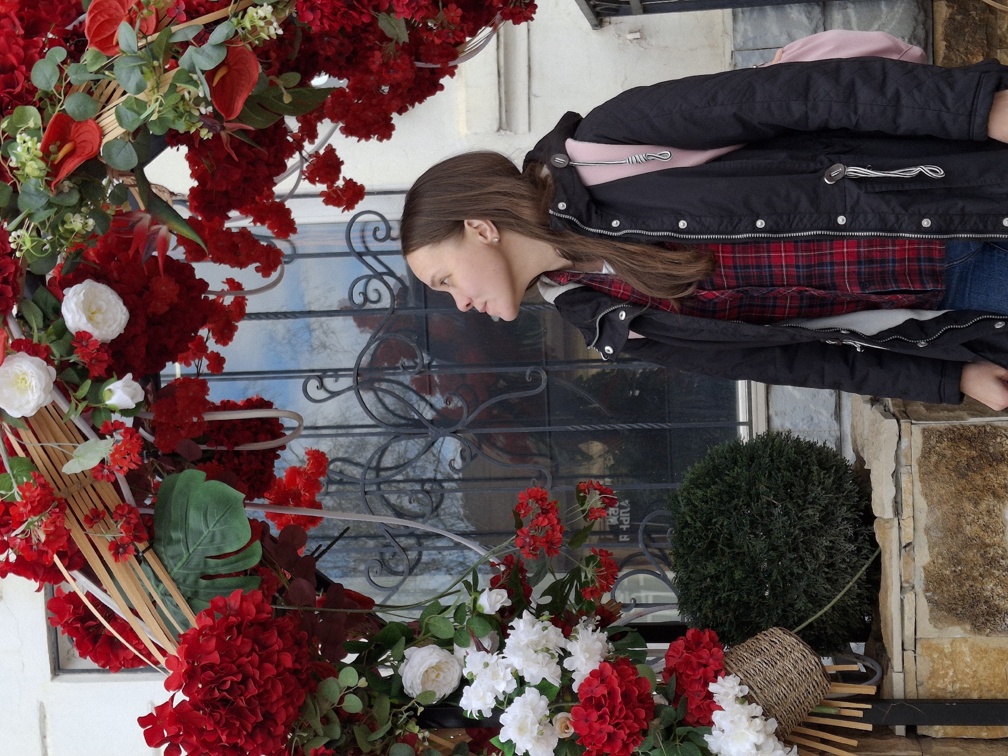A young woman stands in profile next to a vibrant floral display on what appears to be a street or building entrance in Sarapul, Russia. She has long brown hair and wears a black padded jacket over a red and black plaid shirt, blue jeans, and carries a light pink backpack. Her gaze is directed downwards, and her expression is neutral.
To her left, a large floral arch or arrangement dominates the foreground, bursting with abundant bright red flowers, including what appear to be hydrangeas and anthuriums, interspersed with white roses and other small white blossoms. Lush green foliage, including large tropical-style leaves and slender wooden/bamboo sticks, completes the elaborate decoration.
Behind the woman and the flowers, a window with ornate dark metal scrollwork reflects a bright sky. Below the window and to the right, a small spherical green topiary bush and a woven wicker basket are visible, placed on a stand. The background includes parts of a building facade, featuring both smooth light-colored walls and textured stone sections. The scene takes place during the daytime. Faint, distorted Cyrillic characters are vaguely visible in the window's reflection.