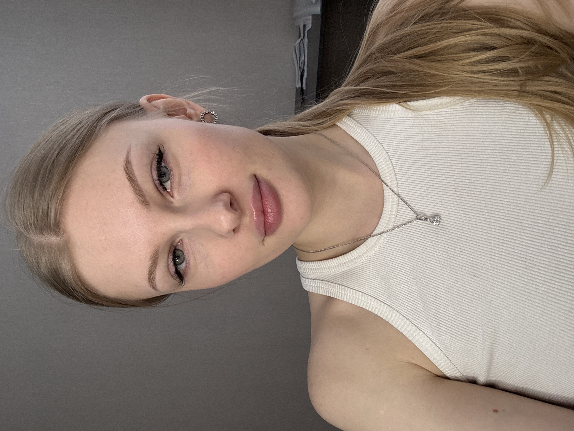 A fair-skinned woman is shown in an indoor setting. She is wearing a white ribbed tank top and a silver necklace with a diamond pendant. Her hair is blonde, parted in the middle, and loosely styled. She has fair eyebrows, eyelashes with noticeable extensions, and pink lips. She is wearing a silver hoop earring in her left ear. The background is a grey wall. It is not possible to identify the location as Perm, Russia from this image.