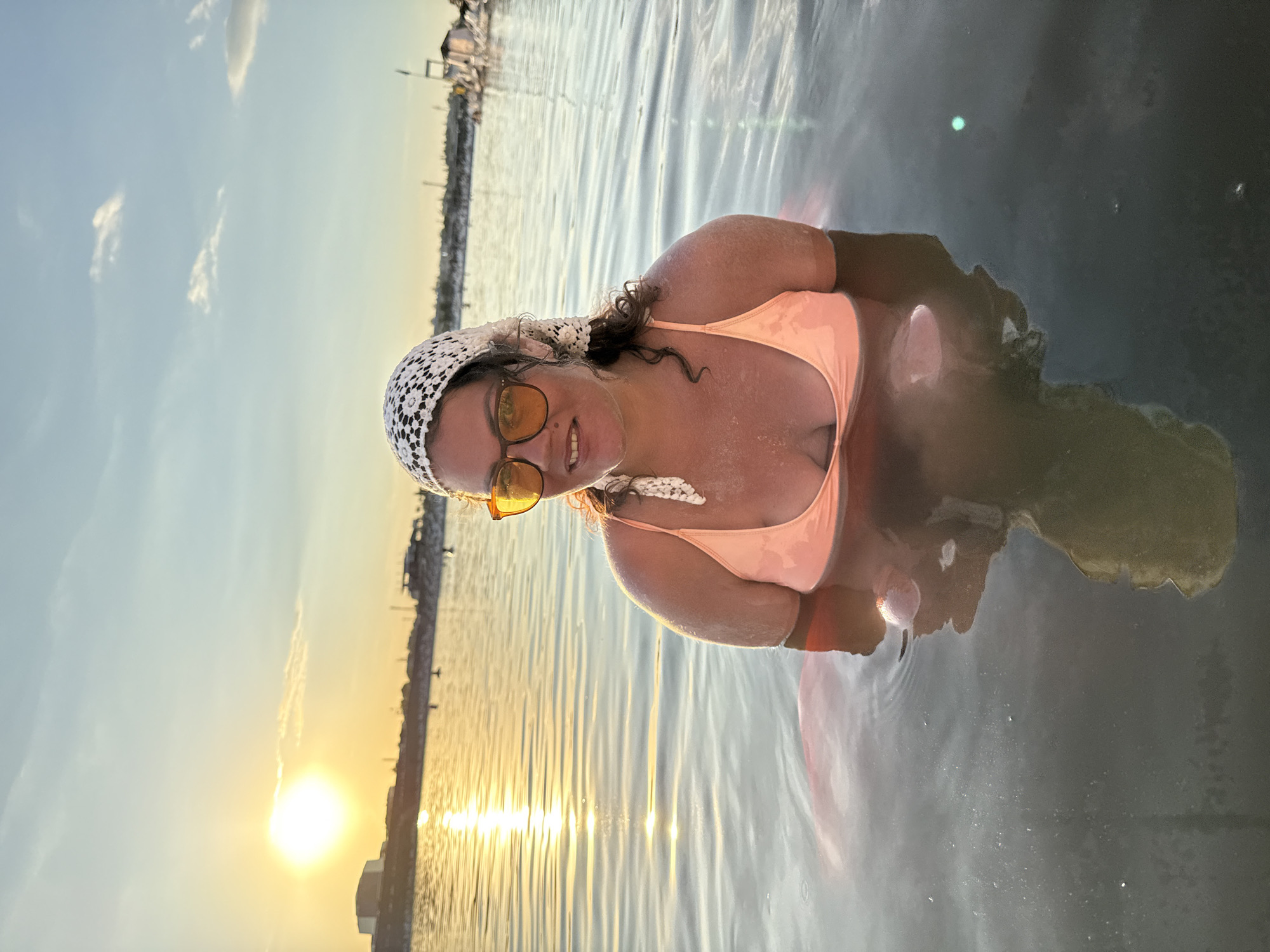 A woman is partially submerged in shallow water, wearing an orange bikini top, a white headscarf, and orange sunglasses. She is looking at the camera and smiling. The water is calm with ripples, and a reflection of the woman and the sky is visible. The background shows a hazy shoreline with buildings and structures, and a bright sun low in the sky, casting a golden reflection on the water. The scene appears to be a natural body of water, likely a salt lake, given the context of Sol-Iletsk, Russia, known for its salt lakes and therapeutic mud. The overall atmosphere is peaceful and serene, with the golden hour lighting enhancing the natural beauty of the location.