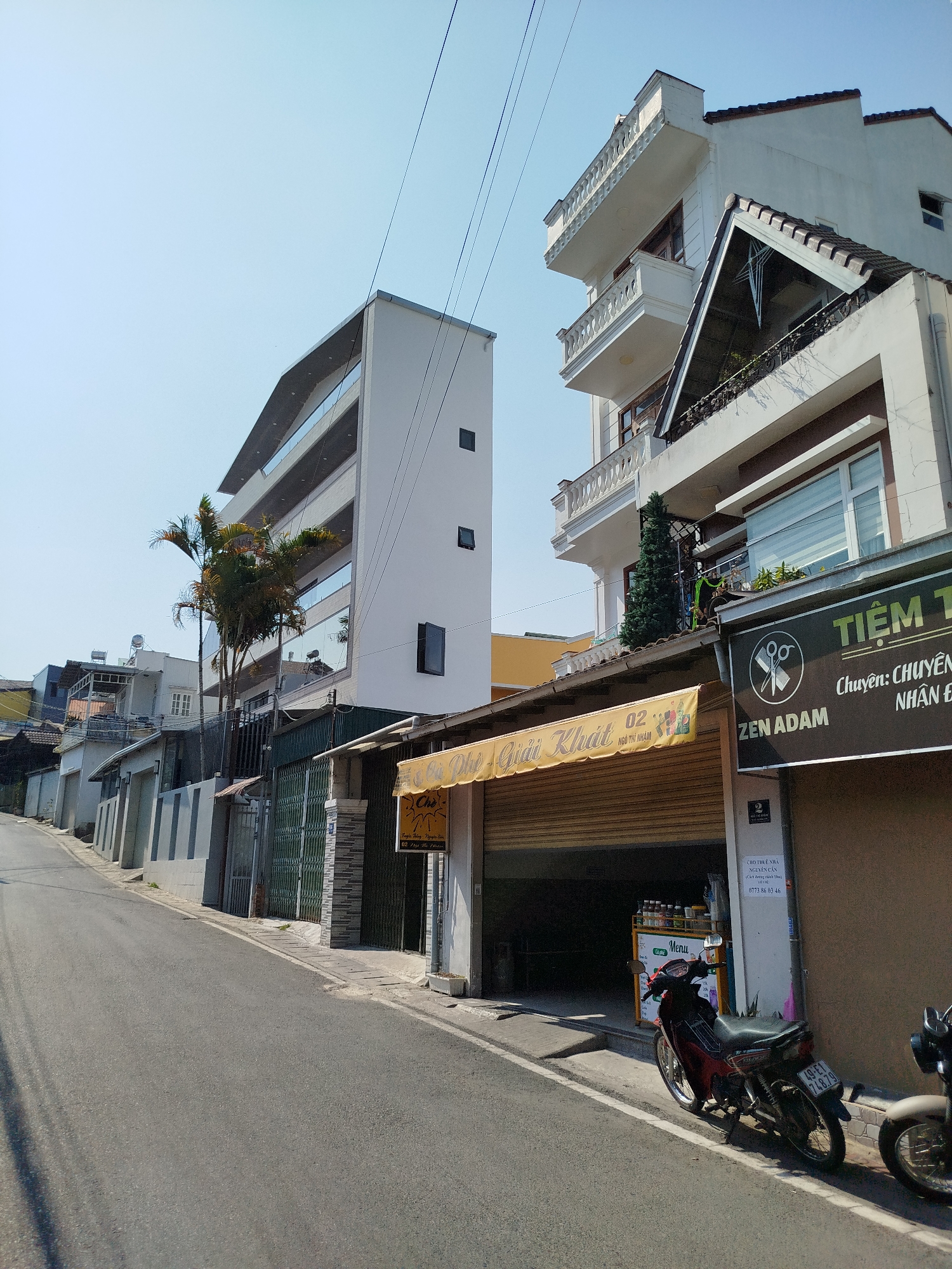 A street in Da Lat, Vietnam, shows a view uphill on a clear day. The street is asphalt and devoid of people. Along the right side of the street is a building with several businesses. The first storefront has a sign reading "Tiệm T - Chuyên: Chuyên Nhận Đ" and "ZEN ADAM" above an illustration of scissors. Adjacent to this is another storefront with a yellow awning labeled "Chè Giải Khát 02 NGÔ THỊ NHẬM" in Vietnamese. A red motorcycle with the license plate "49-E1 748.79" is parked on the sidewalk in front of this business. Next to the motorcycle, also on the sidewalk, is a gray scooter. To the left, on the other side of the street, are houses and fences that line the sidewalk as the road ascends. In the background are more houses and buildings higher up the hill. Overhead, power lines stretch across the sky.