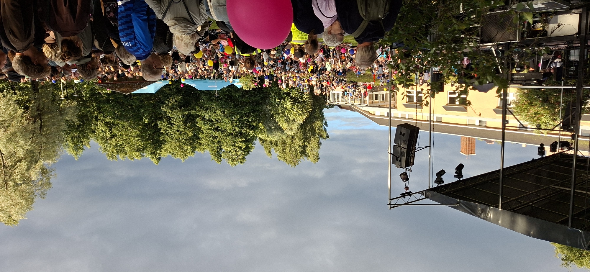This is a wide-angle, outdoor photograph taken at an event in Tuusula, Finland, likely in the summer or early autumn, given the lush green trees and bright, though partly cloudy, sky. The scene is dominated by a large crowd gathered on a grassy area in front of a yellow building and a stage.
On the left side of the image, a black stage structure is visible, with lights and speakers hanging from it. In the background, the yellow building appears to be a community center or school, with large windows and a covered walkway. The crowd is spread out on the lawn, with people sitting and standing, facing the stage. Many are holding or are near colorful balloons, adding a festive atmosphere. Some individuals are wearing bright clothing, including a bright yellow vest worn by a person in the foreground.
A dense line of trees forms the backdrop to the event, with their green leaves catching the sunlight, contrasting with the darker, overcast sky. Several blue tents or canopies are visible among the trees, possibly serving as information booths or vendor stalls.
The overall impression is of a community gathering or festival, with a performance or presentation taking place on the stage. The presence of a large number of people, the stage setup, and the relaxed atmosphere suggest a public event.