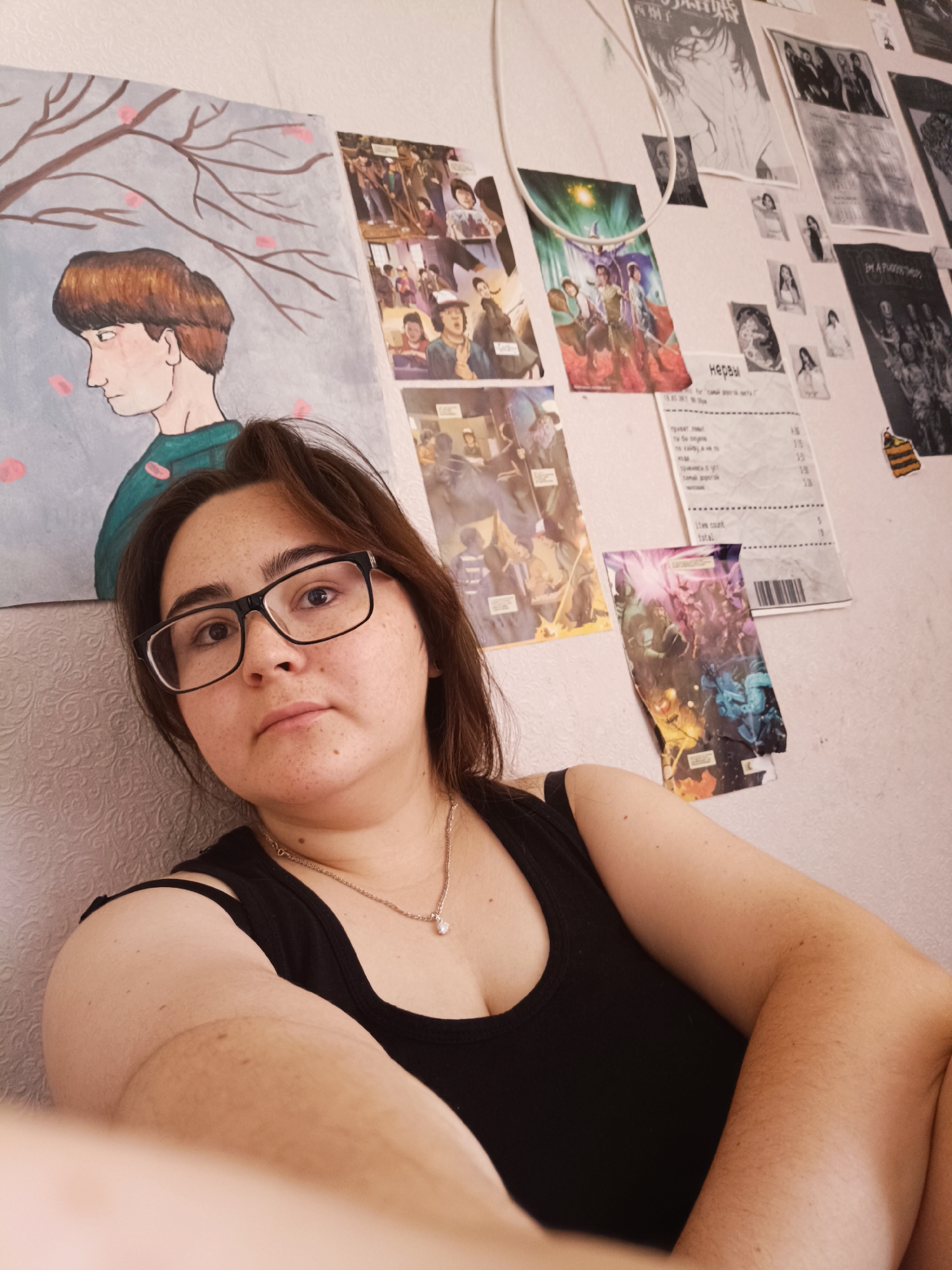 This is a medium shot, eye-level, indoor selfie of a young woman in a black tank top and glasses. She has brown hair and is looking directly at the camera with a neutral expression. The background is a textured wall with various posters and a painting. To the left is a painting of a person with brown hair and a green shirt, with branches and pink blossoms. Several posters, some in black and white, and a crumpled receipt are visible on the wall behind her. The lighting is soft and diffused, suggesting an indoor setting, possibly a bedroom, with no discernible city-specific elements or clues about the location in Посад, Russia.