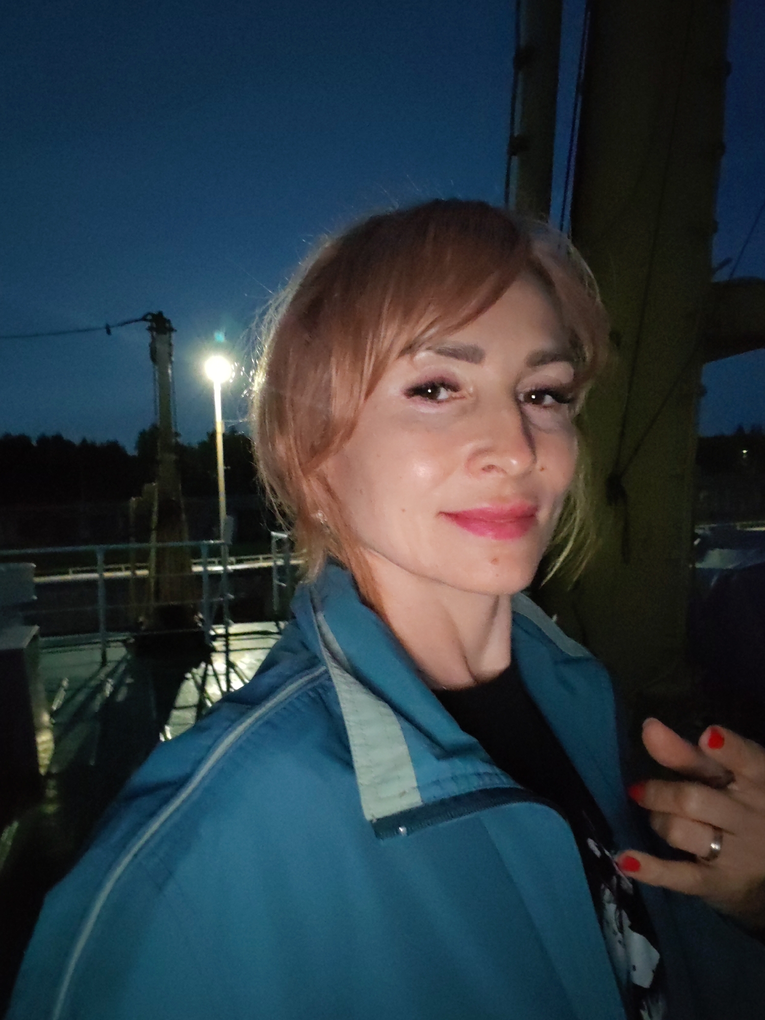 A woman with blonde hair and pink lipstick poses for a selfie at dusk in Sheksna, Russia. She wears a blue jacket over a black shirt. Her right hand, with red nail polish, is visible on the right side of the frame. In the background, a lamppost illuminates a dark sky and a railing along what appears to be a waterfront or dock.