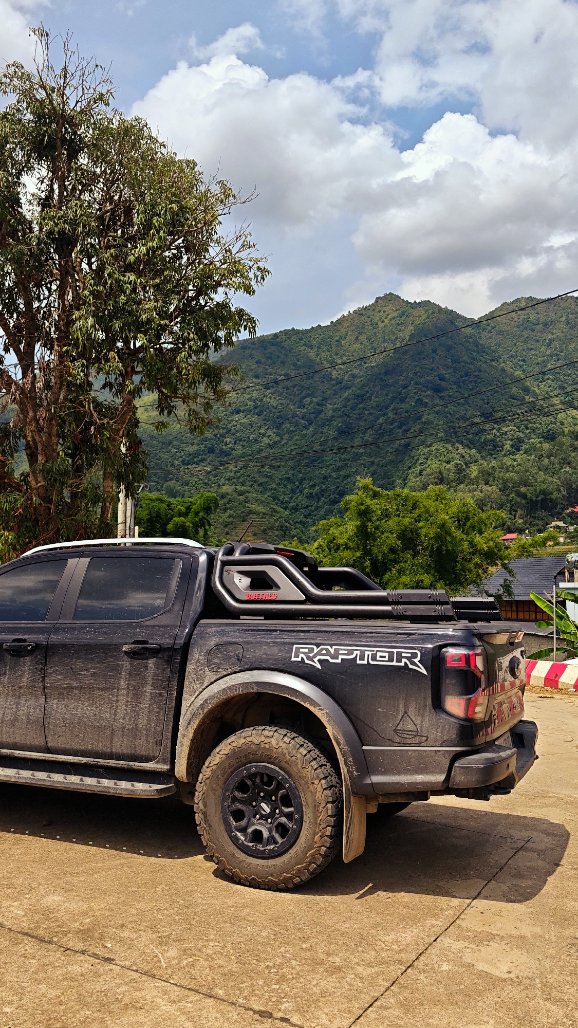 The image presents a daytime view of a mountainous landscape in Mường Kim, Vietnam. A heavily mud-splattered, black Ford Ranger Raptor pickup truck is the central subject, parked on a light brown surface, likely concrete or dirt. The truck is equipped with aggressive off-road BFGoodrich All-Terrain T/A tires and a prominent aftermarket sports bar on its bed, which has "BUNKERED" partially visible on it. Large white "RAPTOR" lettering is emblazoned on the truck's side, and "RANGER" is faintly visible on the tailgate.
To the left, a large, leafy tree partially obscures the background. The backdrop is dominated by lush, densely forested mountains, rising under a bright, partly cloudy sky featuring white and grey clouds with patches of blue. Power lines can be seen stretching across the mountain slopes. In the distance, on the lower slopes of the mountains, glimpses of houses suggest a nearby village or settlement. Distinct shadows cast by the truck indicate it is midday. The scene evokes a sense of outdoor adventure and the rugged natural beauty of the region.