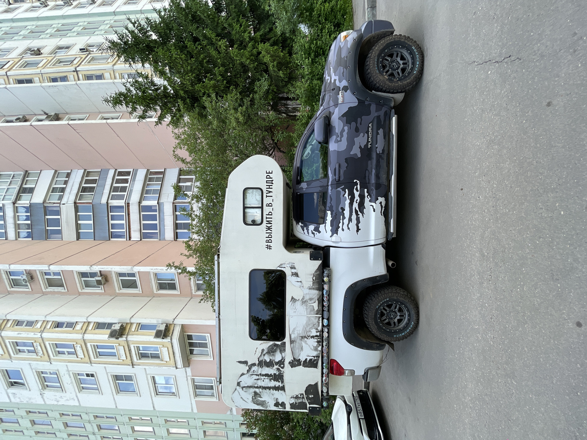 A gray camouflage Toyota Tundra pickup truck with a white camper attached is parked on asphalt next to a multi-story building. The camper has a black and white mountain scene on it and the words "#ВЫЖИТЬ_В_ТУНДРЕ" on the side. The truck is facing to the right of the frame, slightly angled. The building is a light brown and white apartment complex with numerous windows and some air conditioning units. A green tree is visible behind the truck and next to the building. Another vehicle is parked behind the camper. The license plate on the vehicle parked behind the camper is B857XE 797. The scene appears to be located in Moscow, Russia.