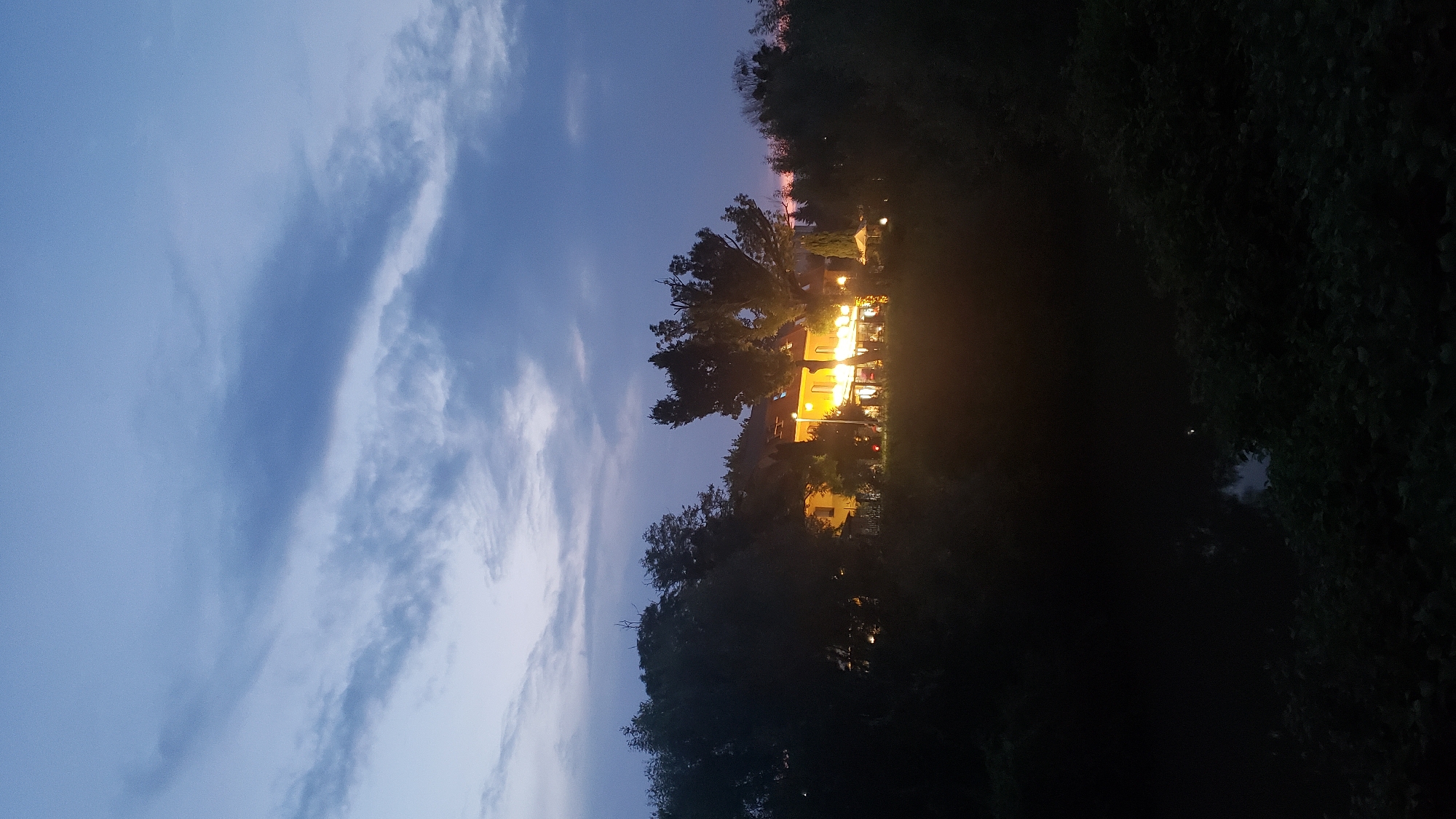 The image captures a twilight scene at a building that appears to be a restaurant or hotel, nestled among trees. The structure is illuminated with warm, yellow lights, indicating it is operational during the evening. The sky above is a deep blue with streaks of darker clouds. The foreground is dominated by dark silhouettes of trees and foliage, with a small, dark body of water visible at the bottom left. The scene suggests an outdoor dining area or a well-lit entrance. This location is identified as the "Lanovka restaurant" in Prešov, Slovakia, which is known for its scenic setting and is situated near a former cable car station.