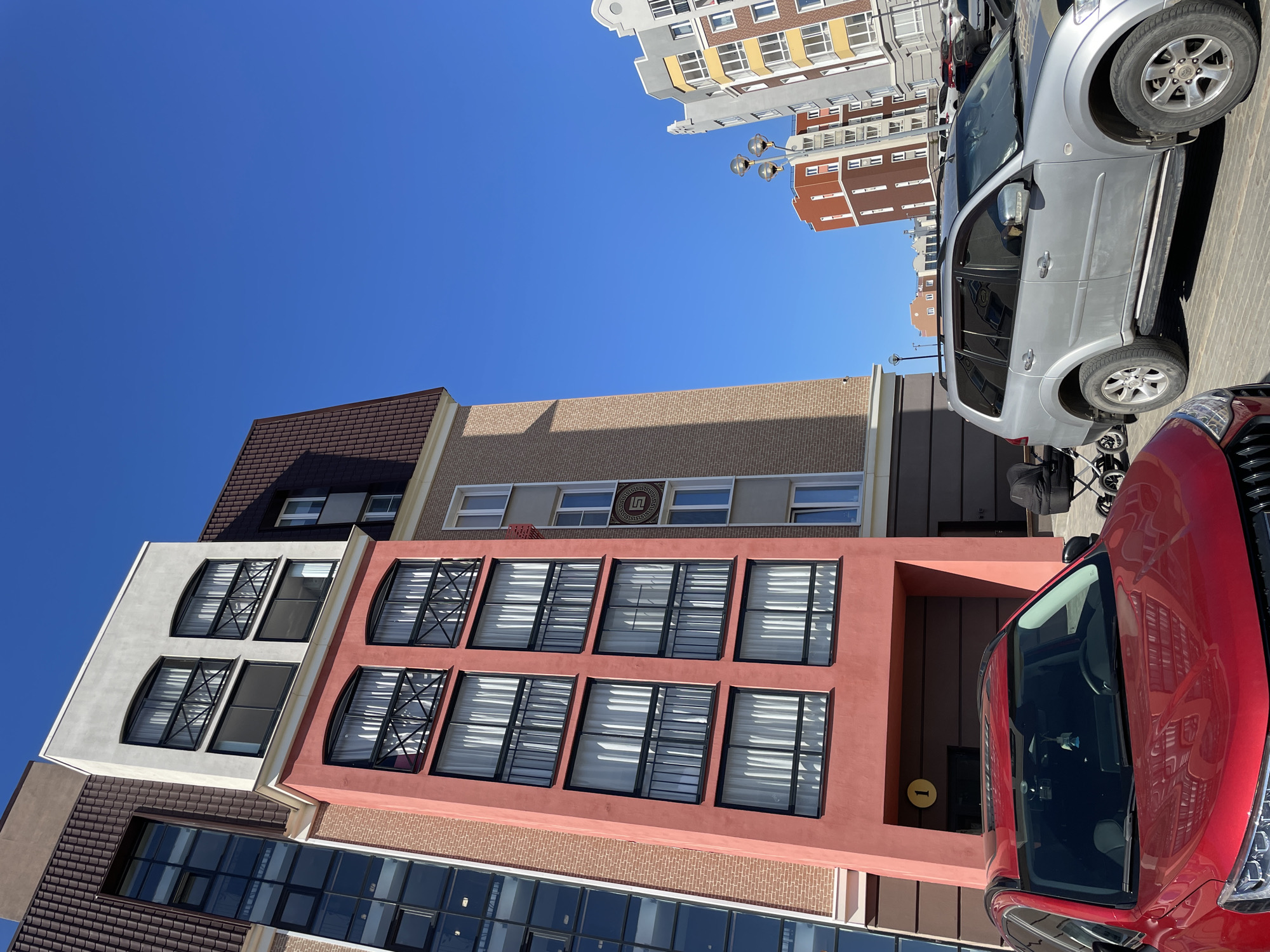 The image shows a view of buildings and vehicles in Pervomaysky, Russia under a clear blue sky. The buildings display varied architectural styles with windows and distinct color schemes. A silver SUV and a red vehicle are parked, partially obstructing the view of the buildings. A street light and a portion of another building can be seen in the background. A stroller is also visible near the silver SUV. A building has windows with black frames and white blinds. A golden circular sign is located beside the entrance of a red building.