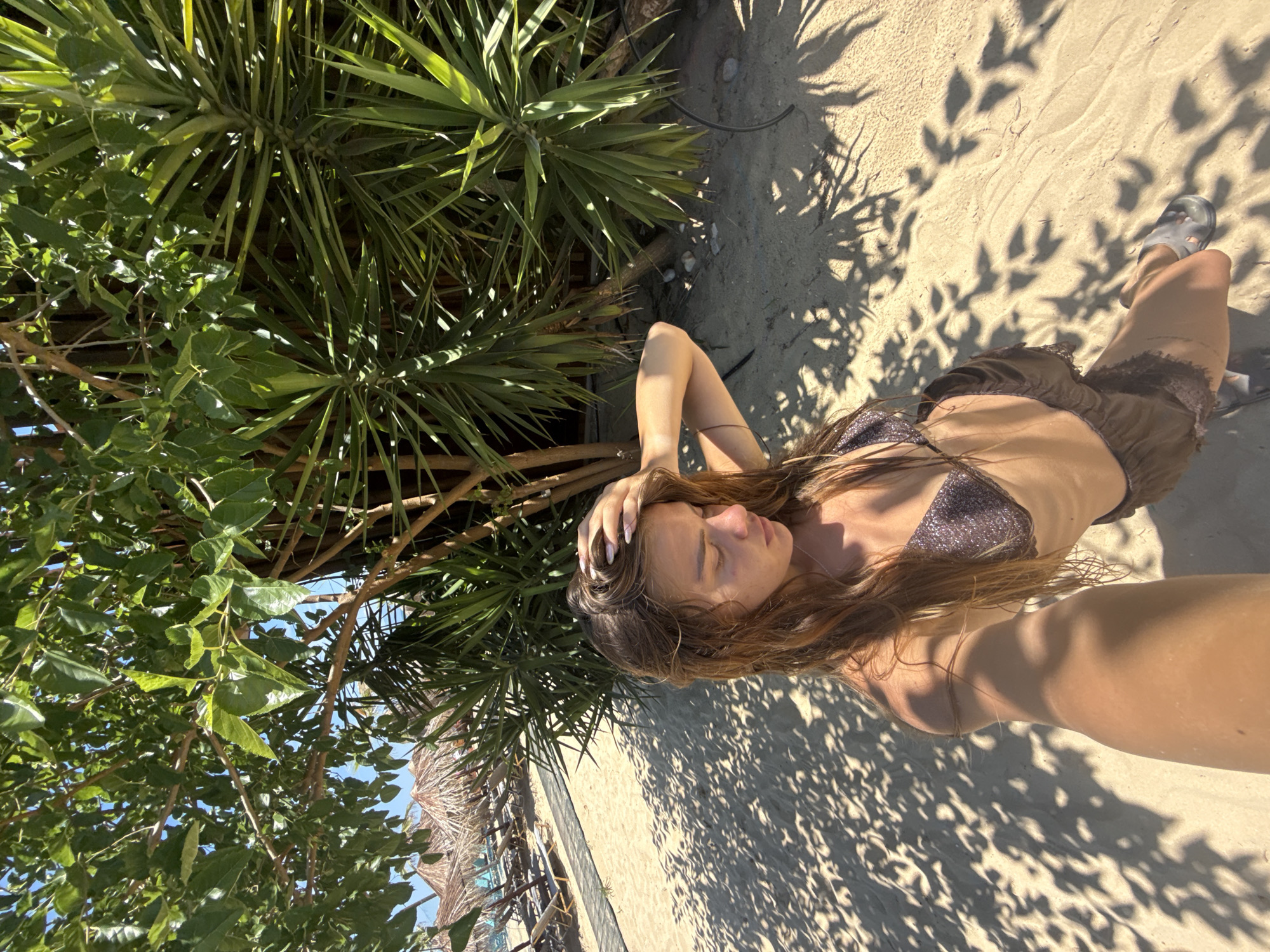 A woman with long, wet, brown hair is taking a selfie on a sandy beach in Nea Peramos, Greece. She is wearing a dark, shimmering bikini top and shorts. Her right hand is raised to her head, and her eyes are closed. Sunlight filters through the leaves of a large tree, casting dappled shadows on the sand. In the background, beach umbrellas and lounge chairs are visible. The environment appears to be a tropical or Mediterranean beach setting, with sandy terrain and lush greenery.