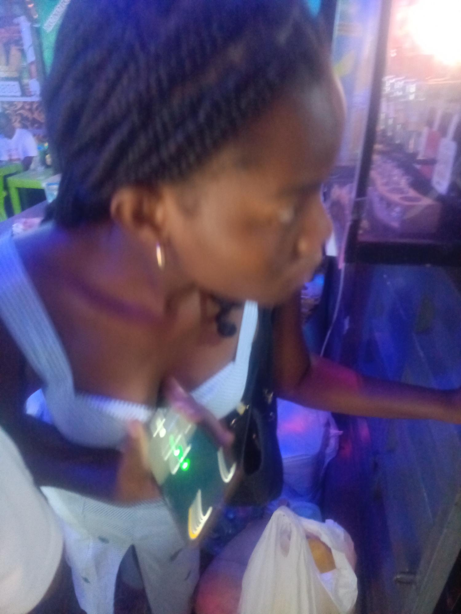 A woman, with dark braided hair, is shown from the chest up in a medium shot in what appears to be a dimly lit interior space with purple and blue lighting. She is wearing a white top with thin straps and is holding a smartphone with a green light on the screen in her right hand. Her left arm is bent upwards. The background is blurry, but suggests a bar or a similar establishment with bottles and shelves visible. The woman's head is turned to the right, and her face is not clearly visible.