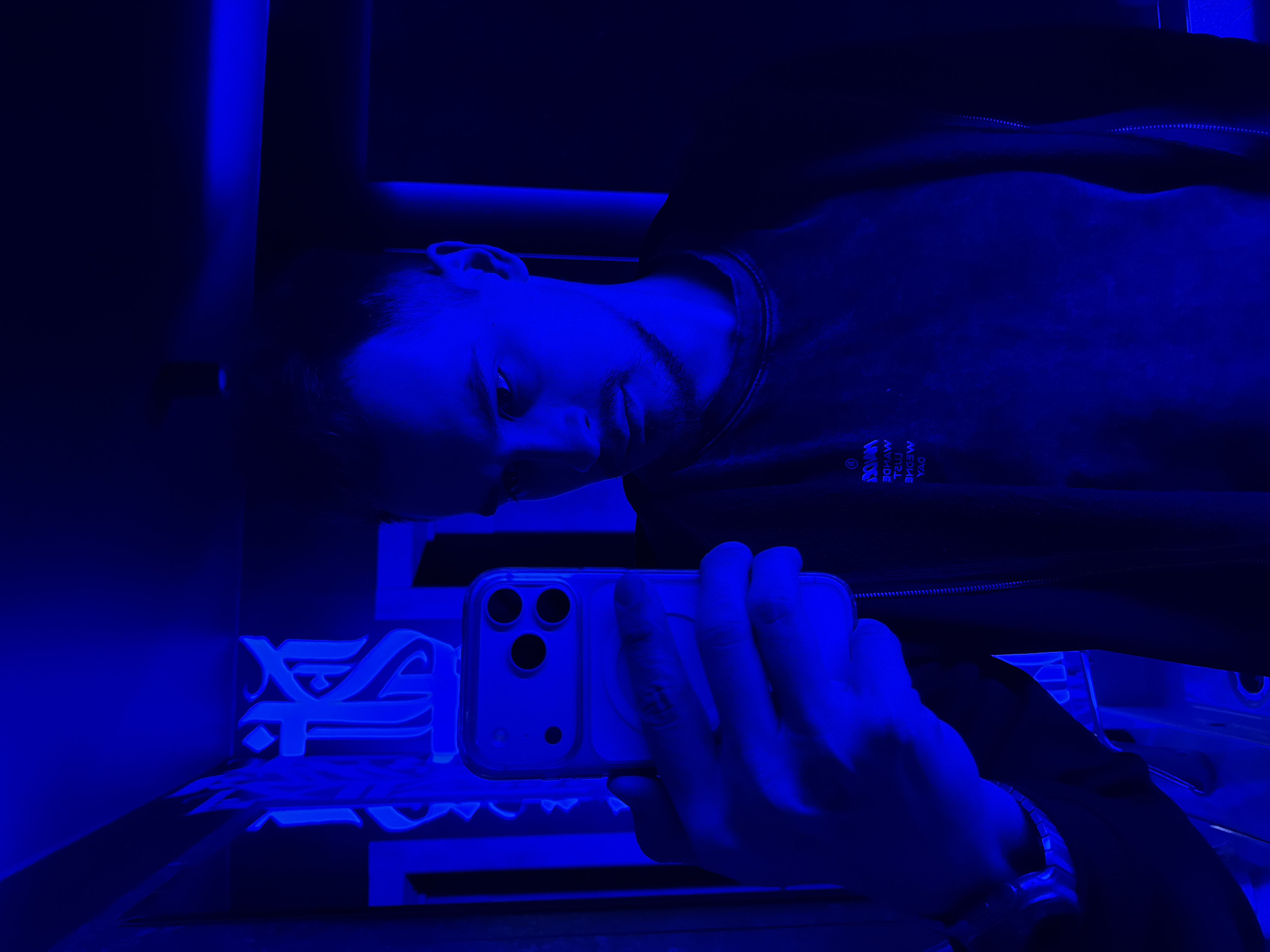 A man is taking a selfie in what appears to be a dark room illuminated by blue light. He is holding a phone with three camera lenses in his hand. The location seems to be a modern or stylized room with some sort of display or signage behind him, possibly an art installation. There is a symbol or logo in the background, which might provide a clue to the location. It's hard to confirm the specific location in Krasnoyarsk based on the image.