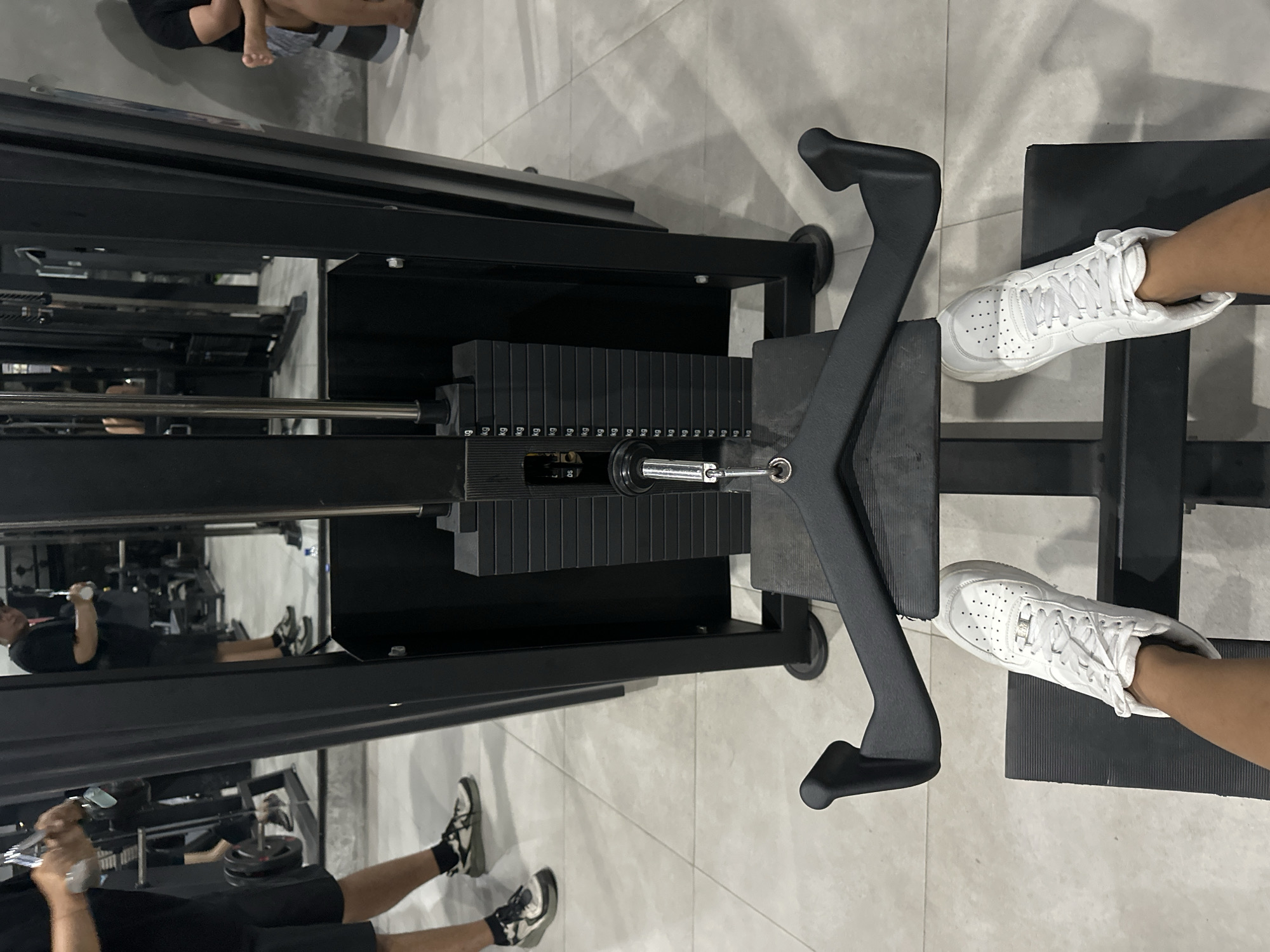 A top-down view shows a leg press machine, inside a gym with marble tiled floors. The machine is black with a vertical stack of weights, and a foot platform. A person wearing white sneakers is positioned on the platform. The background includes other gym equipment and individuals. The location is possibly in Batubulan, Indonesia.
