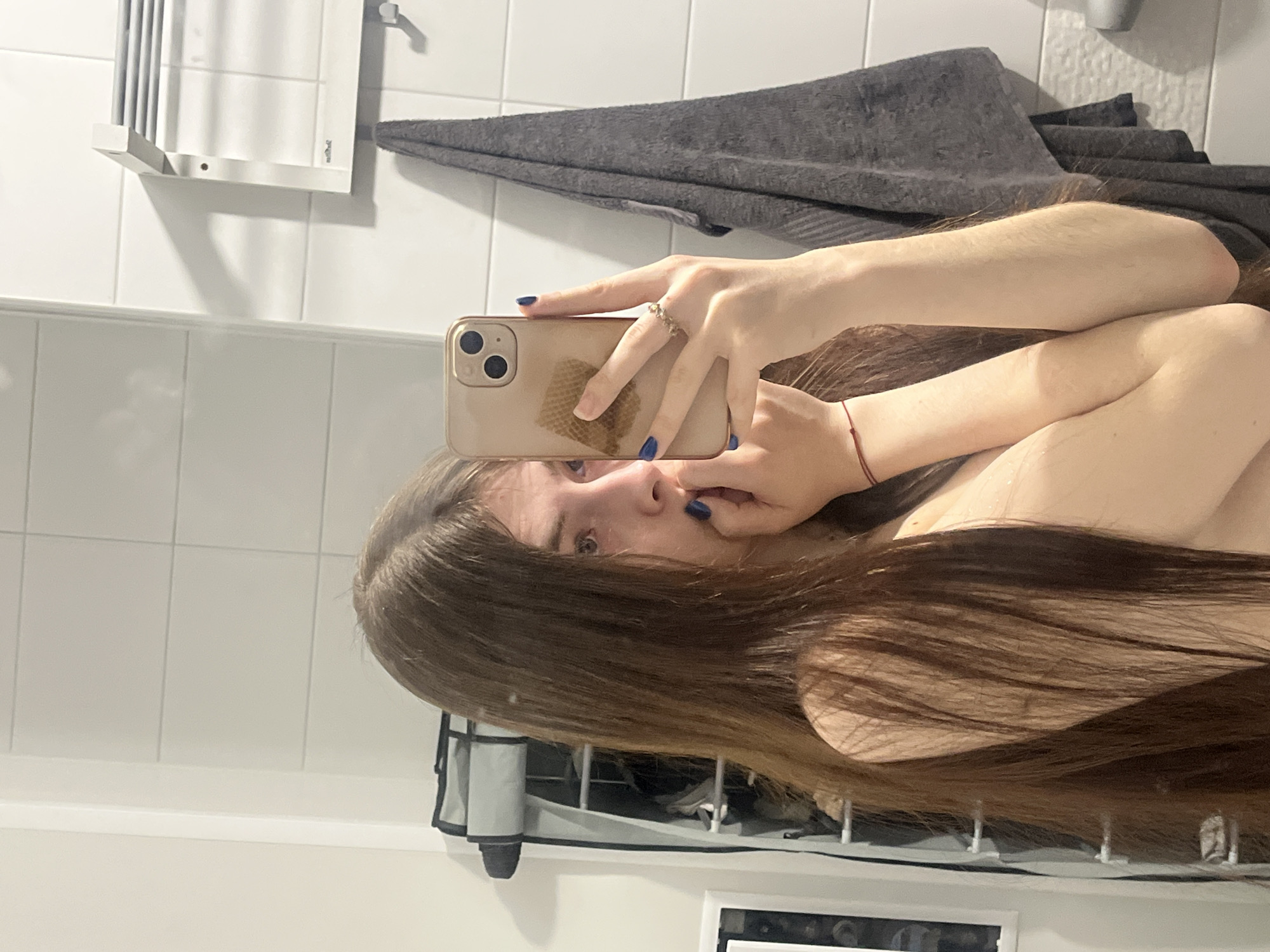 A woman is taking a selfie in a bathroom. She is holding a gold iPhone, and her blue nails are visible. She has long brown hair. The background includes white tiled walls, a gray towel, and a white heated towel rack. The location is not evident from the image.