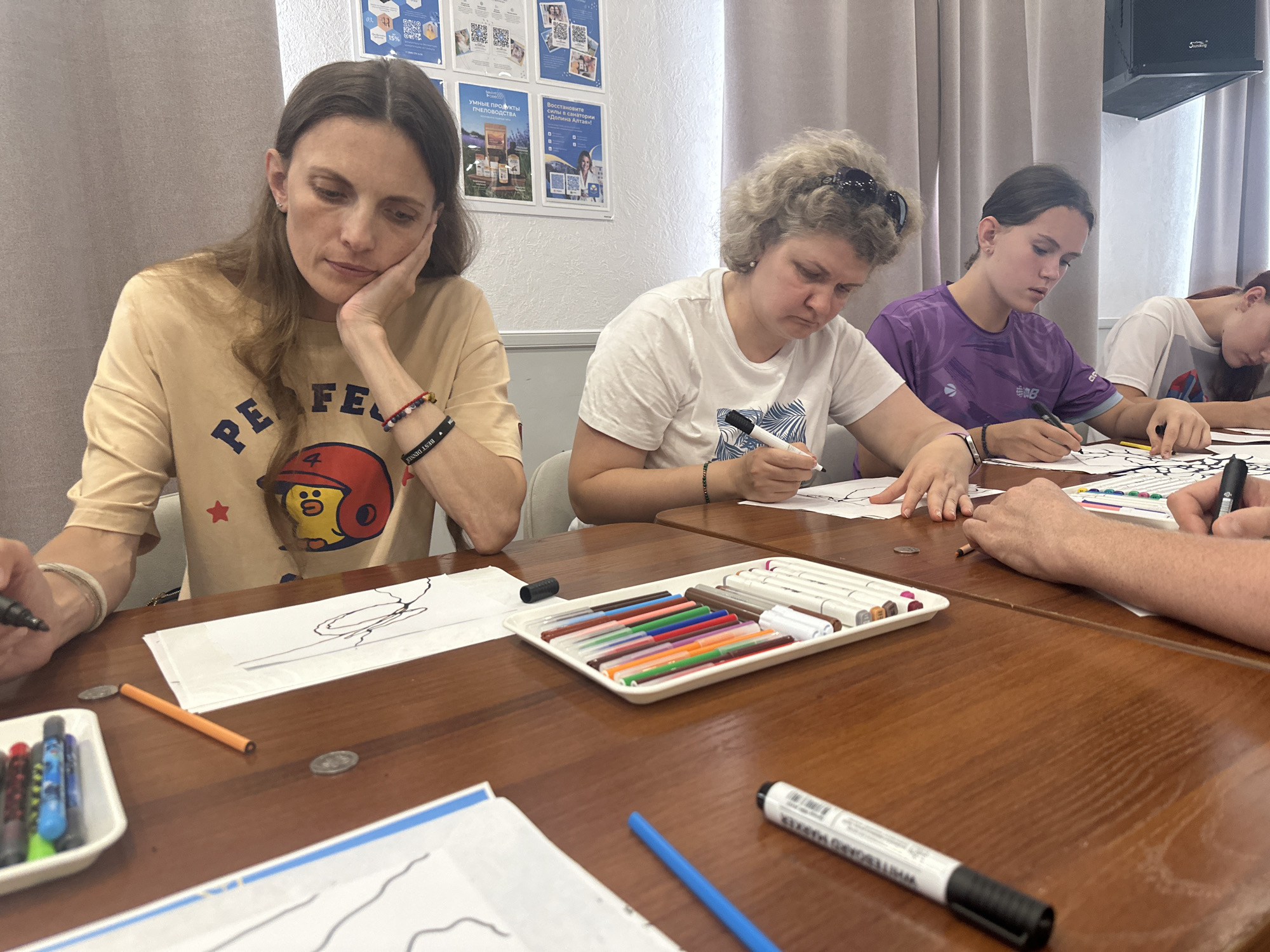 The image shows a group of people, appearing to be women and girls, seated at a wooden table, engaged in drawing or art activity. There are at least four individuals visible, with some partially obscured. They are holding markers and drawing on paper. Various art supplies, including a tray of colorful markers, individual markers, and coins, are spread across the table. The background includes a wall with posters and curtains, suggesting an indoor setting. There is no visual information that directly indicates the location in the city of Даниловка (алтайский), Russia, but the scene depicts a creative or educational gathering.