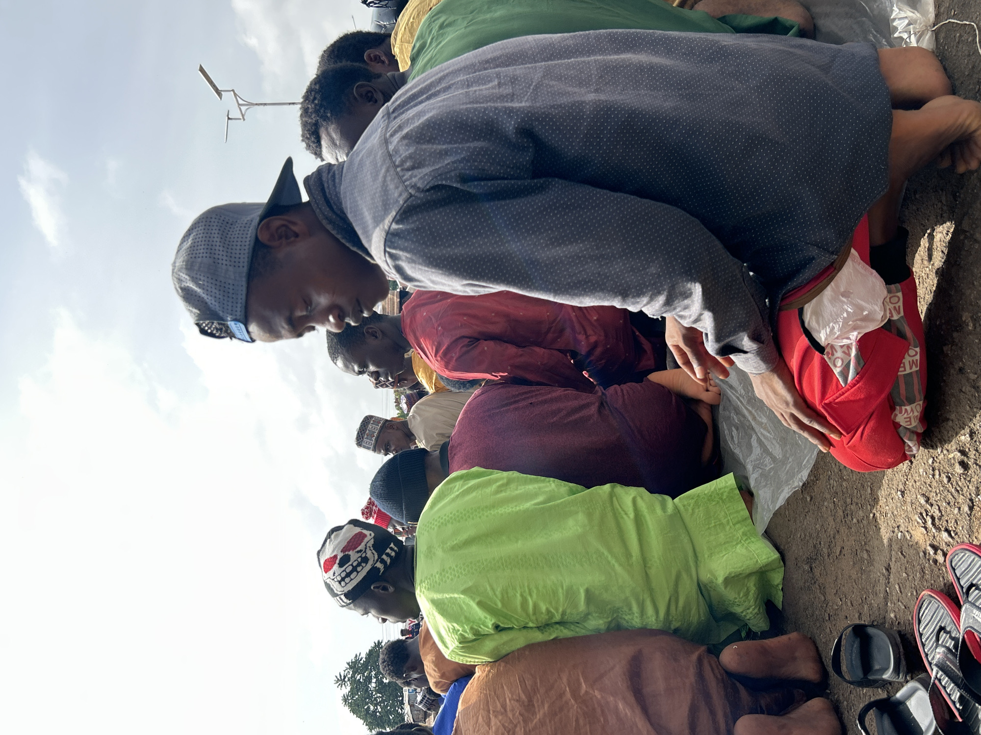 A group of African men are kneeling in an outdoor space, likely a public area, in Jos, Nigeria. They are all oriented in the same direction, their heads bowed in what appears to be a unified action. The men are wearing a variety of clothing, including shirts in shades of green, red, brown, and grey, as well as head coverings such as caps and kufis. They are kneeling on what appears to be a dirt or paved surface. Various pairs of sandals and shoes are placed nearby. Overhead, the sky is visible, and a pole with a mounted light fixture is seen.