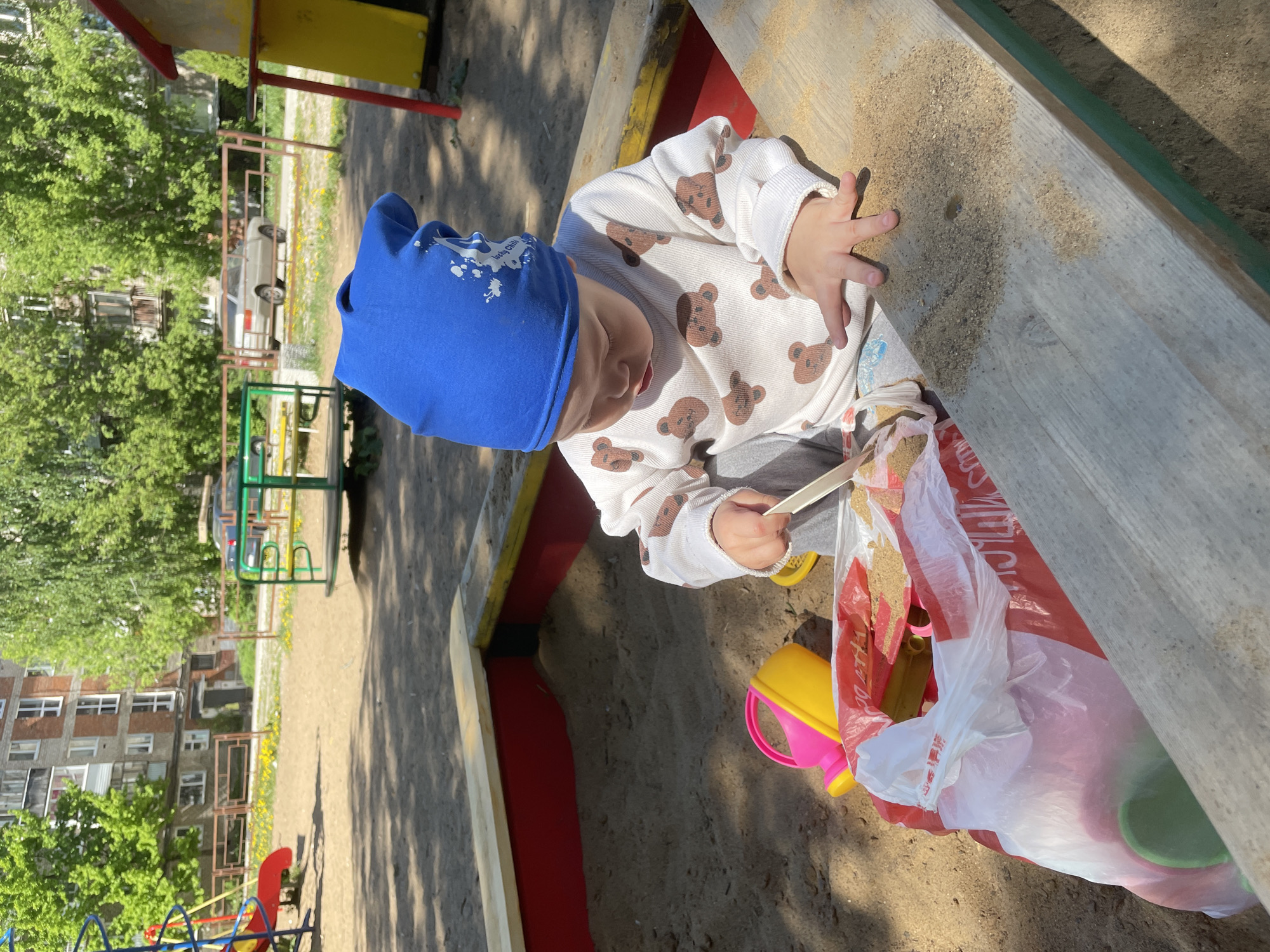 A child in a blue hat and teddy bear-patterned shirt plays in a sandbox. The child is positioned near a wooden edge of the sandbox. A plastic bag filled with sand, a pink and yellow toy bucket, and other toys are present. The sandbox is located outdoors, likely in Perm, Russia, given the prompt's specification. In the background, there is a playground structure, a multi-story building, and some greenery.