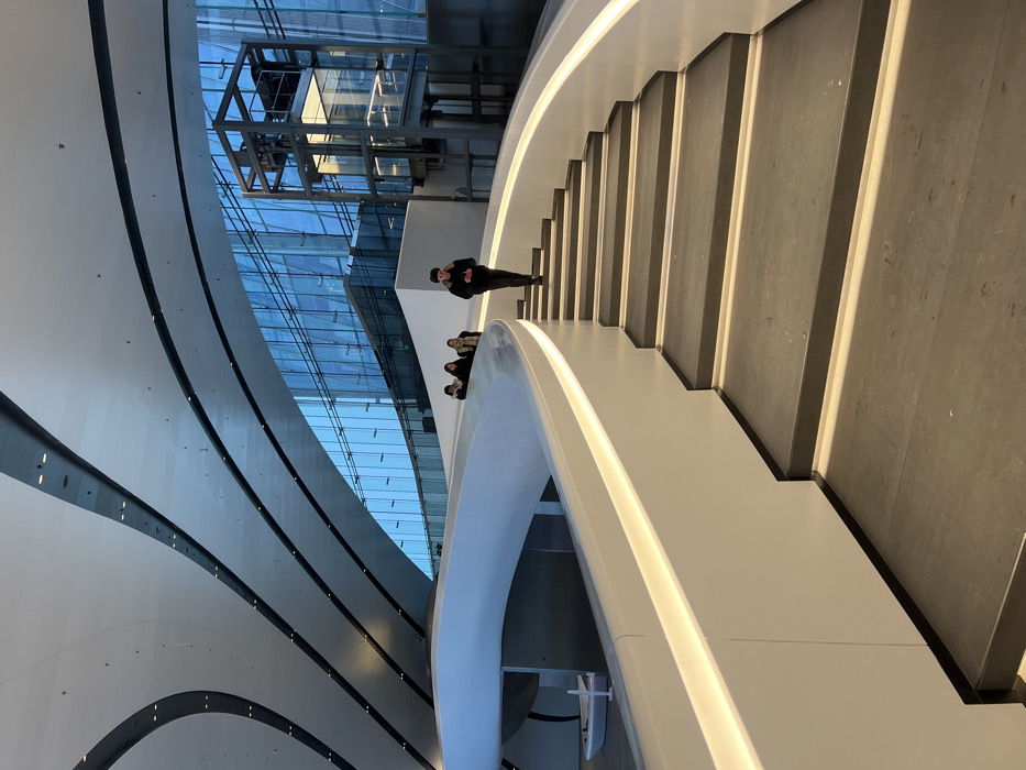 The scene is set within the modern, curvilinear interior of the Lakhta Center in Saint Petersburg, Russia. A wide, dark grey staircase with illuminated risers descends from the right foreground, bordered on the left by a curving white balustrade featuring a continuous horizontal light strip. Parallel to the staircase on the left, a broad, white, illuminated ramp or walkway curves upwards.
Four individuals are present. One young male, attired in a dark jacket, dark pants, and a dark beanie, is descending the staircase, looking down at a mobile phone held in his hands. Further back on the ascending ramp, three additional individuals, appearing to be young adults dressed in winter coats and scarves, are walking away from the viewer.
The background features expansive, curved glass curtain walls, reflecting a cool blue light and structures beyond. Through the glass on the right, a multi-story, open-frame metallic structure housing an elevator shaft is visible. The ceiling consists of multiple curved white panels with integrated linear dark accents and recessed lighting. A small white object, resembling a miniature vessel or sculpture, is located on a lower level beneath the curving ramp on the far left. The interior is brightly illuminated by integrated architectural lighting and ambient light from the exterior.