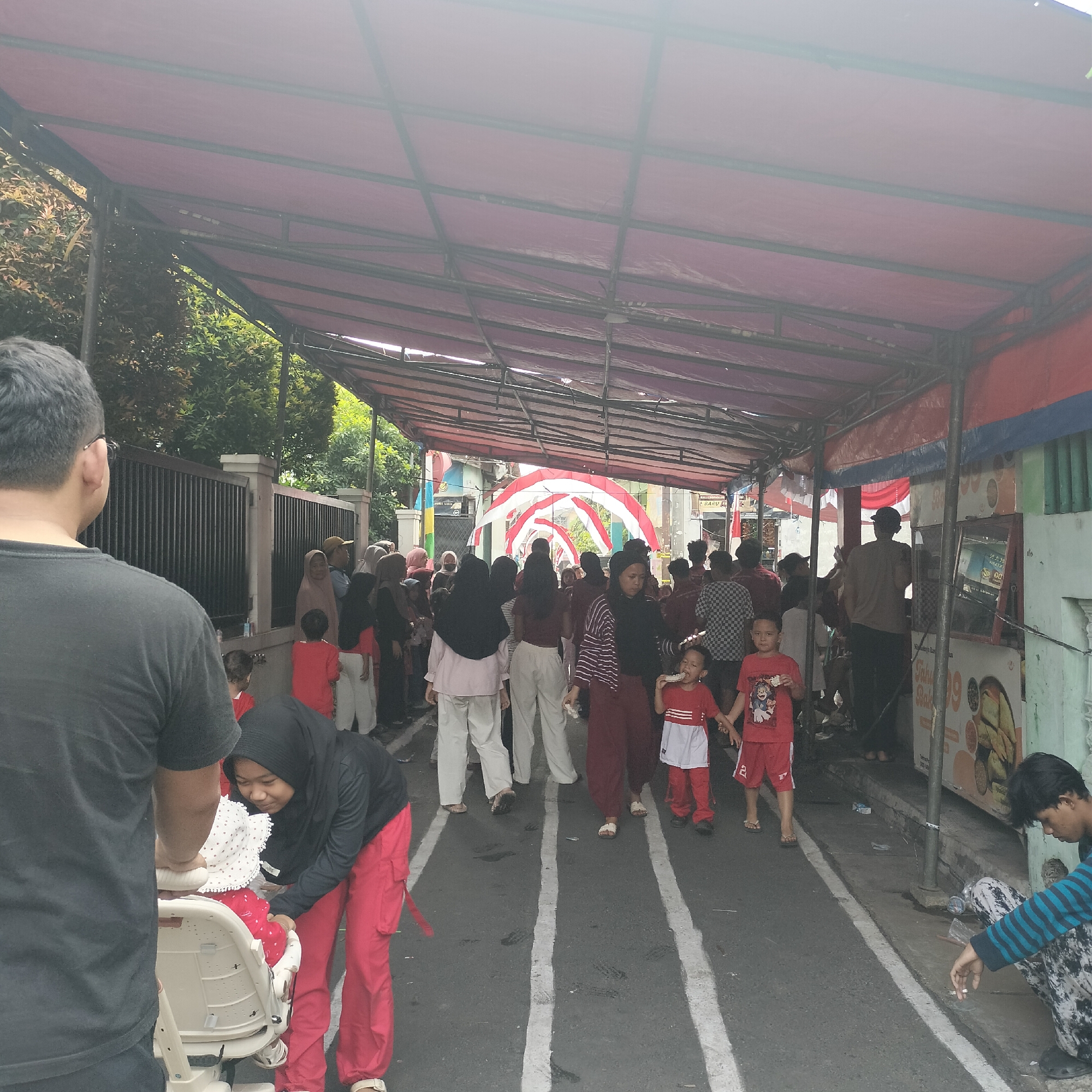The image depicts an outdoor street scene in Tangerang, Indonesia, possibly during a celebration or event. A temporary canopy, supported by metal poles, covers a street area, with a red tarp forming the roof. Underneath the canopy, a crowd of people, including adults and children, are gathered. Many individuals are dressed in red and white clothing, suggesting a patriotic or festive theme, perhaps related to Indonesian Independence Day.
In the foreground, on the left, a man with short dark hair and glasses is visible from behind, wearing a black t-shirt. To his right, a woman wearing a black hijab and red pants is attending to a child in a white stroller or carrier, which is decorated with a white and red polka-dot pattern. The child is wearing red clothing.
Further into the scene, a line of people are walking along a street marked with white lines. Some individuals in the middle ground are seen talking and interacting. There's a decorative arch in the background made of red and white fabric, resembling the Indonesian flag colors, which adds to the festive atmosphere.
On the right side of the image, there are market stalls or food vendors. One stall has a mirror and signage. A young man in a striped shirt and patterned pants is sitting on the curb, smoking a cigarette. Several other people are standing or walking near the stalls. The environment includes buildings with green and light-colored facades, as well as lush green trees bordering the area. The overall impression is a lively gathering in a public space.