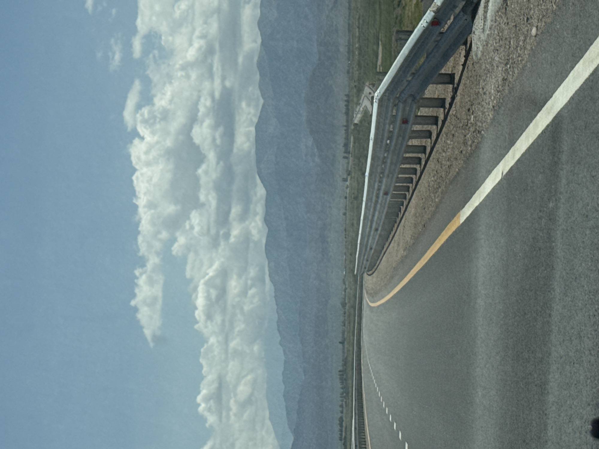 This is a photograph taken from a moving vehicle on a highway. The highway is paved and has lane markings. A metal guardrail runs along the right side of the road. In the background, there are mountains and a vast expanse of green, grassy terrain. The sky is blue with scattered white clouds. The image appears to have been taken during the daytime under bright sunlight, which casts shadows on the road. The overall scene depicts a rural or mountainous landscape, characteristic of areas surrounding Balykchy, Kyrgyzstan. There are no people or specific landmarks visible in the image that would definitively place it within the city of Balykchy itself, but the environment is consistent with the region.