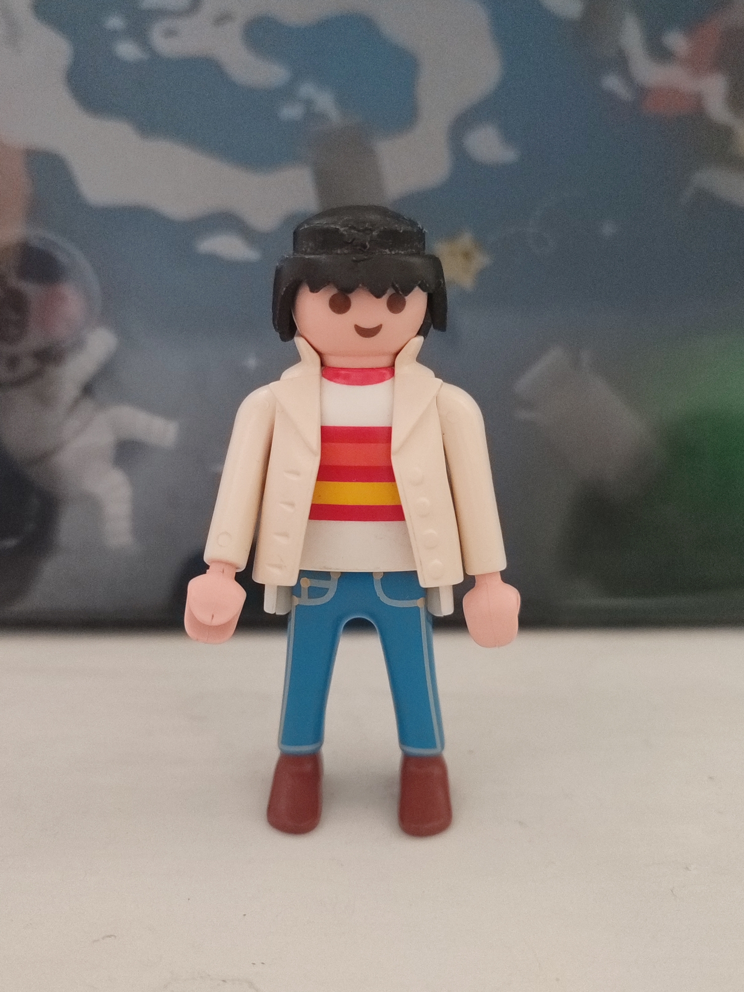 A Playmobil figure of a person with black hair and a smiling face stands in the center of the frame. The figure is wearing a white jacket over a striped shirt with horizontal bands of red, orange, and yellow. The figure's pants are blue, and its shoes are brown. The figure is standing on a light-colored surface. In the background, there is a blurry image that appears to be a map or a mural with shades of blue and white. To the left of the figure, there is a faint, blurred image of what looks like an astronaut in a spacesuit. To the right of the figure, there is a blurry green shape. The lighting is diffused, and the focus is on the Playmobil figure. There is no indication of the city of Chișinău, Moldova in the image.