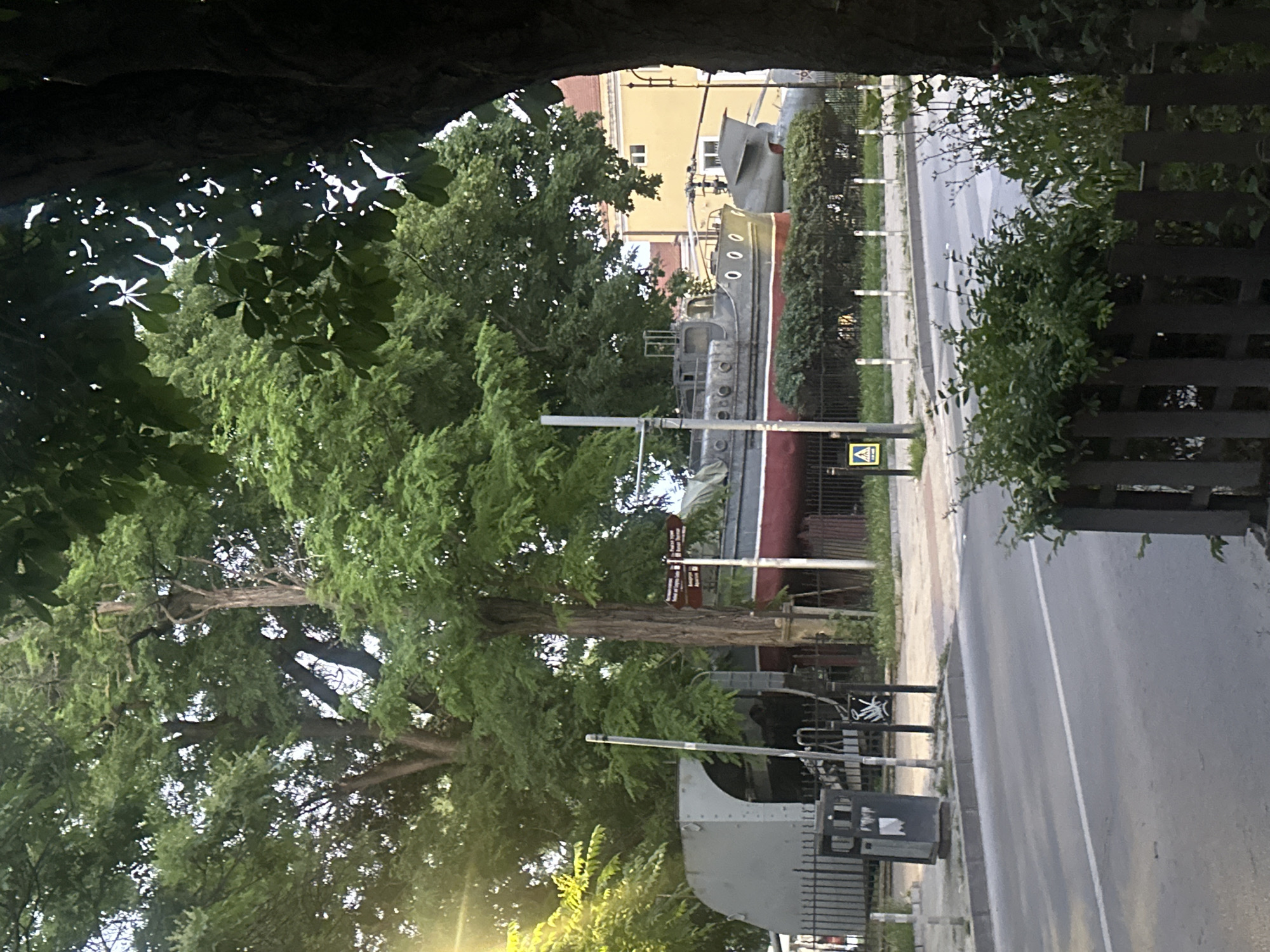 A grey, red, and white boat is on display outdoors, partially obscured by trees and foliage. The boat appears to be a museum exhibit. A road runs in front of the display. There are trash cans and fencing around the exhibit. A yellow building is visible in the background. The time of day appears to be late afternoon or early evening.