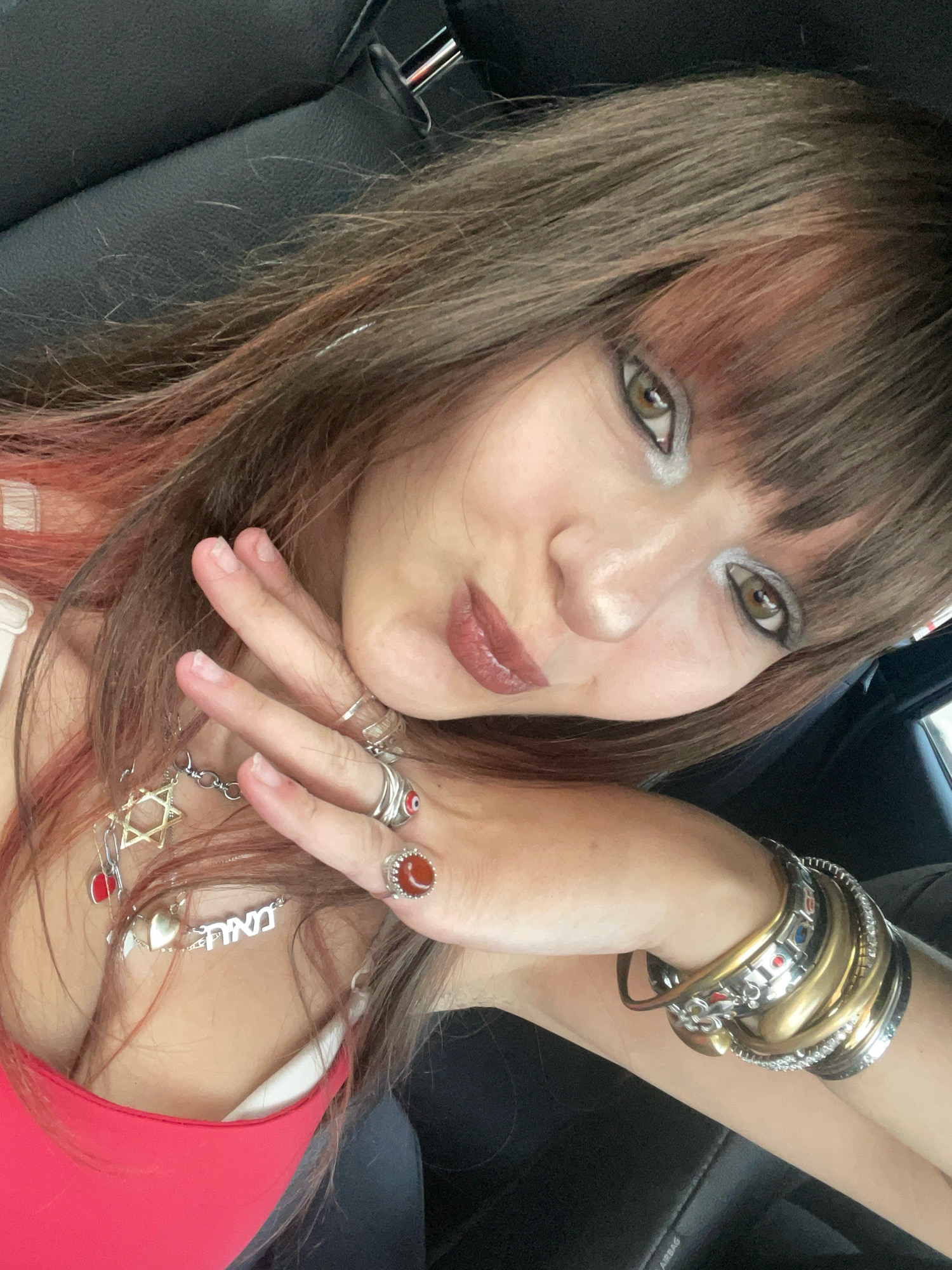 The image is a close-up selfie of a woman with long, brown hair with pink highlights, taken from a slightly elevated angle inside a vehicle. She is wearing makeup, including eyeshadow and lipstick, and several pieces of jewelry: a Star of David necklace with red charms, a silver necklace with Hebrew lettering, multiple bracelets on her left wrist, and rings on her fingers, one of which appears to be a carnelian stone. Her right hand is positioned near her face, with fingers extended. She is wearing a bright red top. The background shows the interior of a car, with dark upholstery and a headrest visible. The lighting suggests it is daytime. No specific location in Tel-Aviv is discernible from the image.