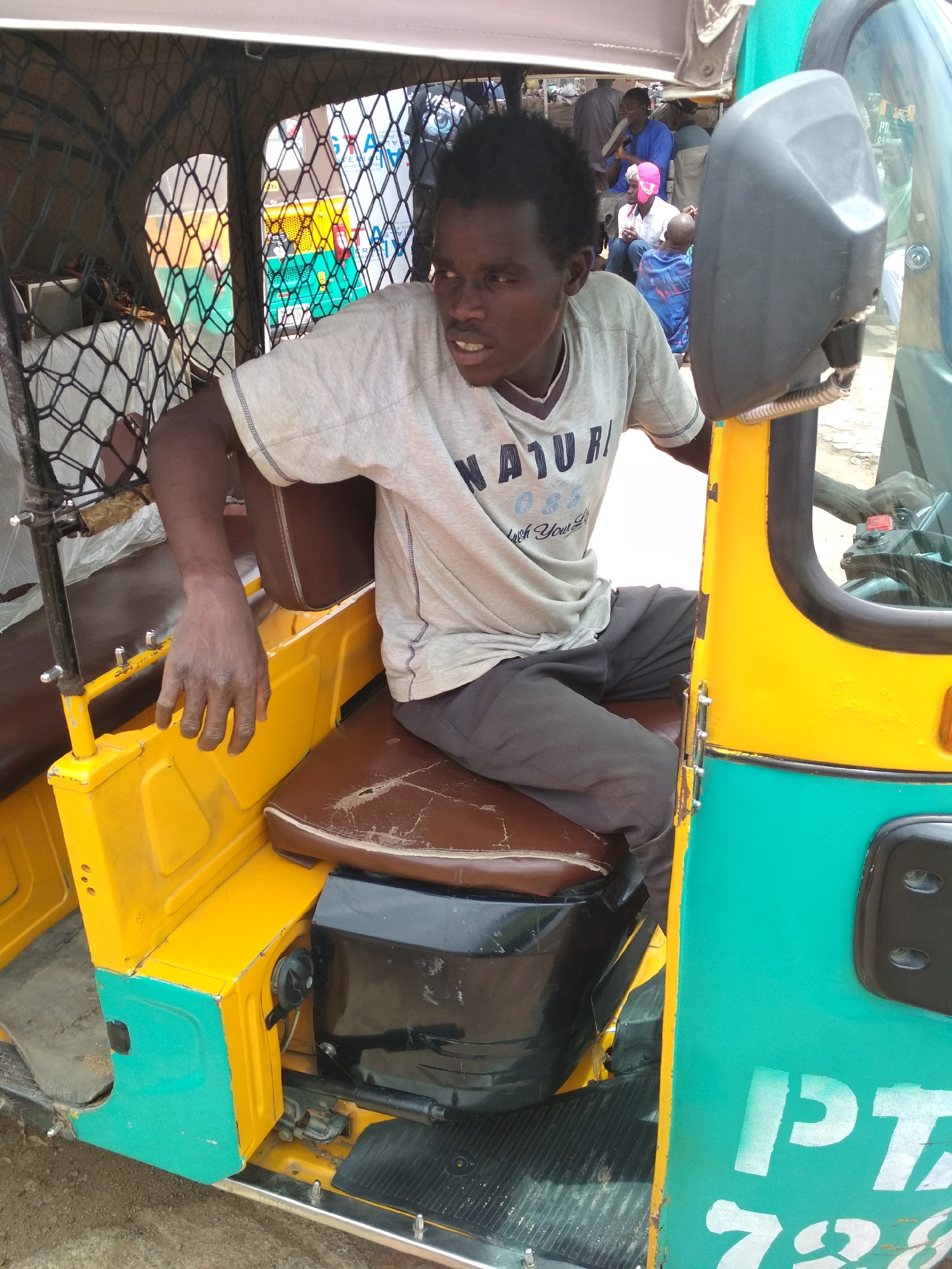 A man with dark skin and short, curly black hair sits in the driver's seat of a three-wheeled vehicle, potentially a motorized tricycle taxi known as a "keke." He wears a light grey t-shirt with dark pants. The vehicle is painted in yellow and teal. The scene is set outdoors, with people visible in the background, possibly waiting for or using transportation. The numbers "728" are partially visible on the vehicle, suggesting it's marked for identification or licensing. The location is within the city of Gura Top, Nigeria.