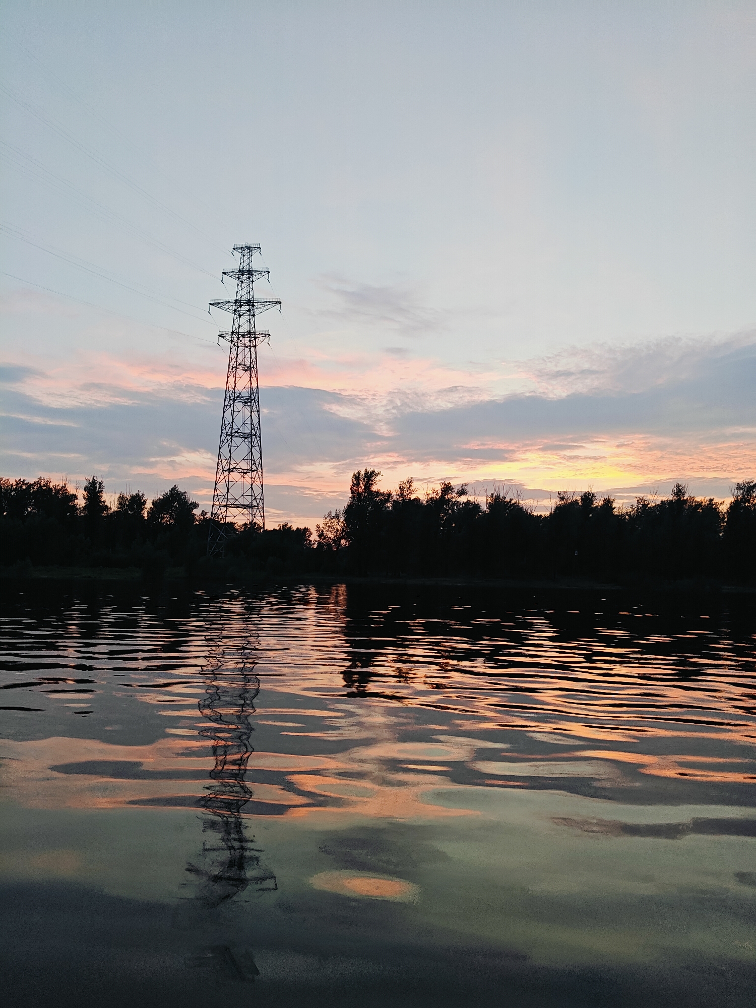 The image depicts a sunset over a body of water with a power line tower and silhouetted trees in the background. The sky displays soft hues of pink, orange, and blue, with scattered clouds. The water reflects the sky and the power line tower, creating rippling patterns of light and shadow. The scene appears to be located in or near Noviy, Russia, given the natural landscape and the presence of power infrastructure. There are no people visible in the image, and the environment suggests a tranquil, natural setting at dusk.