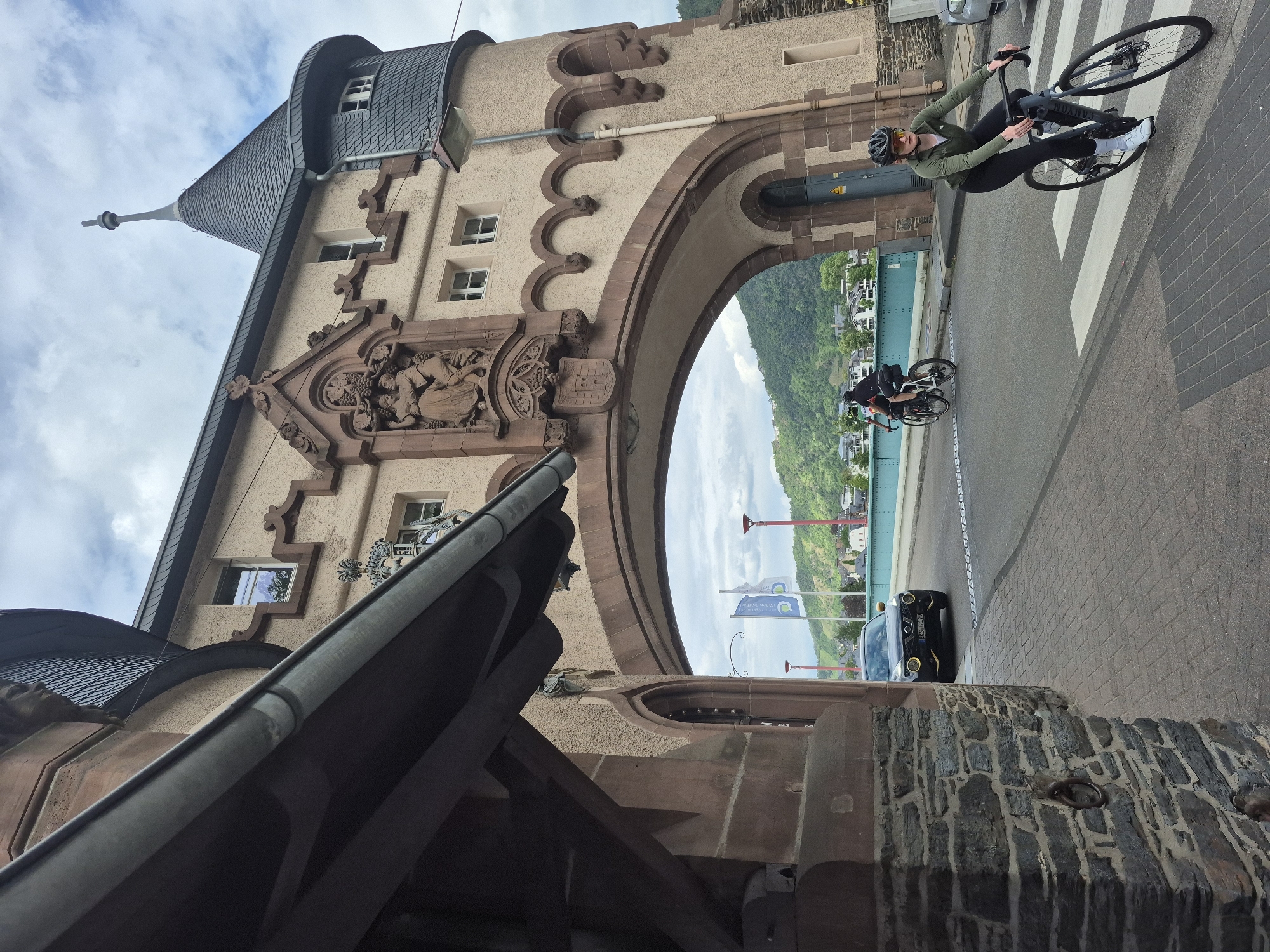 The image depicts a street scene featuring the historic Brückentor (bridge gate) in Bernkastel-Kues, Bernkastel-Wittlich district, Rhineland-Palatinate, Germany, on an overcast day.
In the foreground, a female cyclist, wearing a dark green long-sleeved shirt, black leggings, a black helmet with yellow-tinted sunglasses, and white sneakers, rides a dark grey road bicycle. She is positioned on the right side of a paved road, partially on a white pedestrian crossing. Her gaze is directed slightly to her left. To the far left, a stone pillar or wall is partially visible.
The central feature of the image is the large Brückentor, a multi-story building constructed from light brown stucco with reddish-brown stone accents. It features a prominent arched passage through which the road passes. Above the arch, a detailed relief sculpture depicts two figures, possibly associated with grape harvesting, flanked by decorative elements and surmounted by a pointed gable. Below the sculpture, a carved coat of arms is visible. To the right, a round tower with a conical dark grey slate roof and a finial extends upwards. The building has multiple rectangular windows with white frames. A grey metal gutter is visible along an overhang on the left side of the structure.
A black Nissan Juke car, featuring distinctive yellow circular daytime running lights and yellow trim around its grille, is driving under the archway, moving away from the viewer. Its German license plate, "BKS C 199," is partially visible. Further beyond the car, another cyclist, dressed in dark attire, is visible riding on the bridge.
The road extends onto a bridge with light blue railings, visible behind the Brückentor. On the bridge, red streetlights and two blue and white flags on poles are present. In the background, lush green hills, characteristic of the Moselle valley, rise under a cloudy sky.