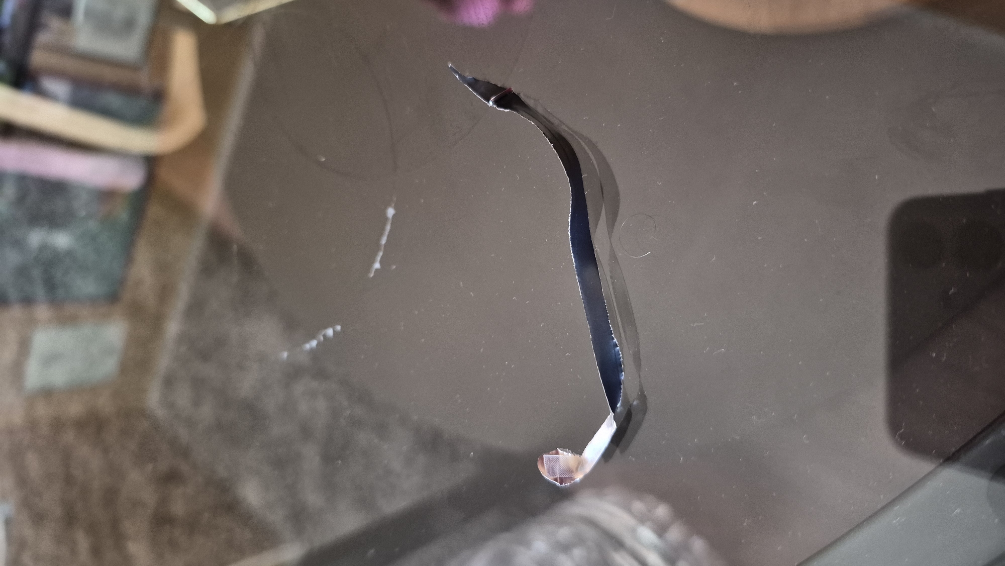 A dark, curved strip of material lies on a reflective, dark surface. The strip appears to be peeling or torn, with one end curling upwards and revealing a lighter, possibly metallic, underside. The surface shows some smudges and dust. In the background, out of focus, are reflections of what appear to be furniture and possibly a wall. The overall impression is that of a close-up shot of an object on a tabletop.