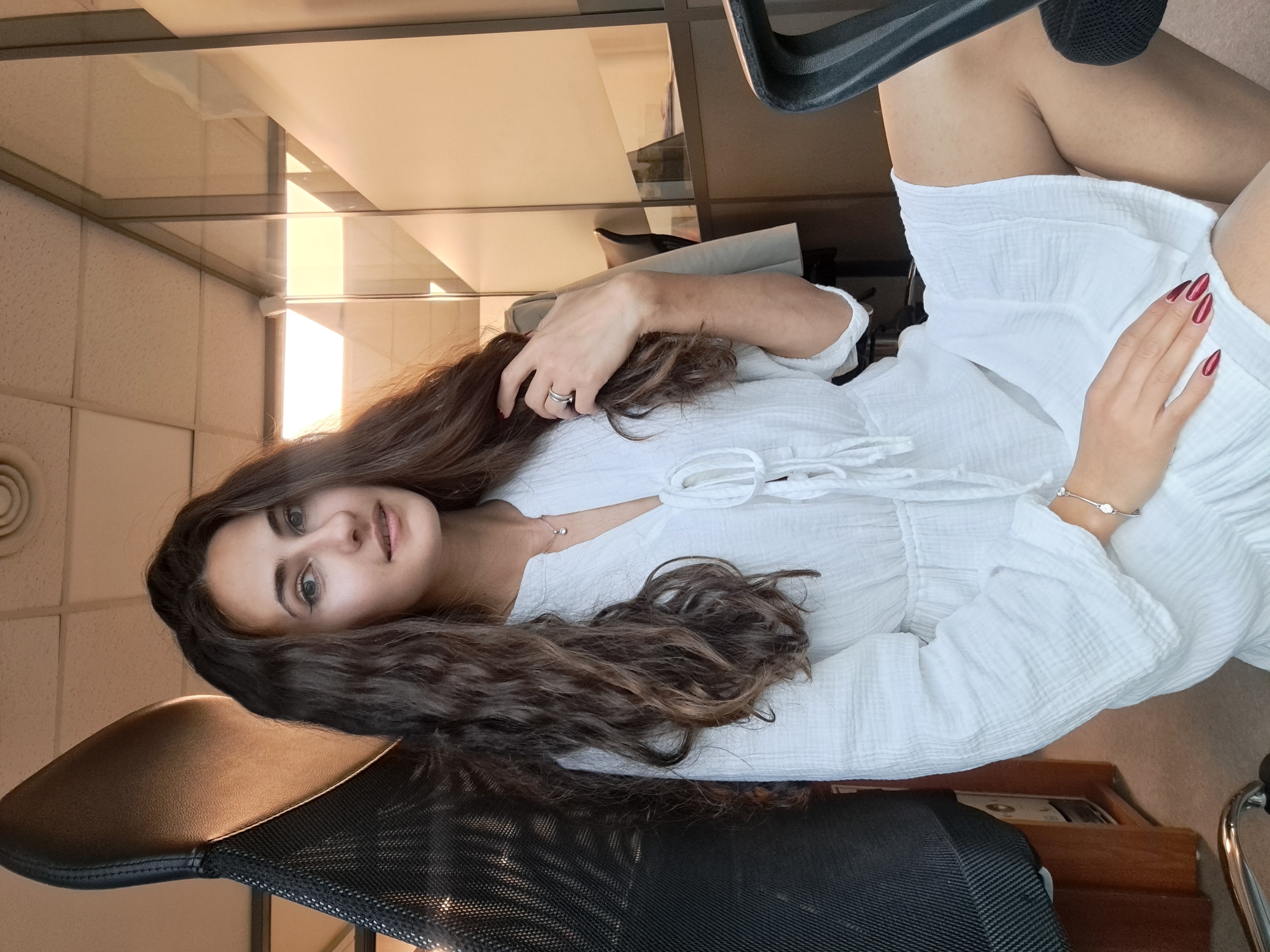 A young woman with long, wavy brown hair sits in an office chair. She is wearing a white dress. Her left hand is resting on her hair, and her right hand is resting on her leg. She is wearing a ring on her left hand and a bracelet on her right. The background shows an office setting with a window and a desk. The city of Sofia, Bulgaria cannot be determined from this image.