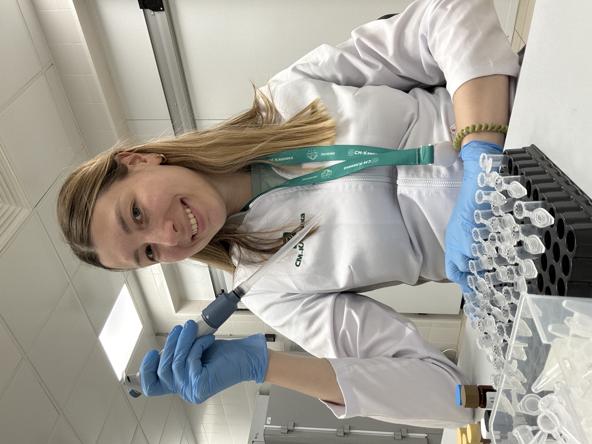 A woman in a white lab coat and blue gloves is working in a laboratory. She is holding a pipette in her gloved hand and appears to be transferring liquid into small tubes that are arranged in a rack. She is smiling at the camera. The lab appears to be modern and well-lit with overhead fluorescent lighting. The woman has a green lanyard around her neck with the words "СМ-Клиника" written on it. This translates to "CM-Clinic", a medical clinic chain located in the city of Moscow, Russia.