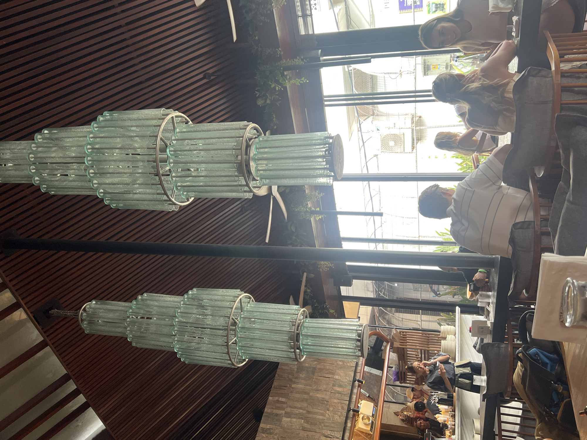 This image depicts the interior of a restaurant or cafe located in North Kuta, Indonesia. The scene is illuminated by natural daylight entering through large windows along the right side.
The ceiling features dark wooden slats arranged diagonally and two prominent, multi-tiered chandeliers. Each chandelier is composed of numerous vertical tubes of light blue-green frosted glass, arranged in diminishing concentric rings from bottom to top, suspended by metal frames. Ceiling fans and hanging green foliage are also visible near the windows.
The back wall on the left side is constructed of stacked, light-colored stone or brick. Below it is a long service counter with a white marble top. Behind this counter, a female staff member wearing a dark grey top and apron is standing, looking at her mobile phone. To her left, another staff member in a white cap is partially visible, facing away from the camera. Stacked white plates and various bottles are on the counter.
In the mid-ground and foreground on the right, several patrons are seated at dark wooden tables with chairs that have dark fabric cushions. A man in a white and blue striped polo shirt is seated with his back to the camera, looking left. Across from him, a woman with long blonde hair, also with her back to the camera, is facing a third woman who has long dark hair, is smiling, and looking left. Another person in dark clothing is partially visible at the same table. On the table, there are drinking glasses, napkins in holders, and a white, skull-shaped decorative object. Bags, including a dark backpack with a blue section and a beige tote bag, are placed on or next to chairs in the foreground.
Beyond the large windows, parts of external buildings, fences, and what appear to be air conditioning units are visible under bright daylight.