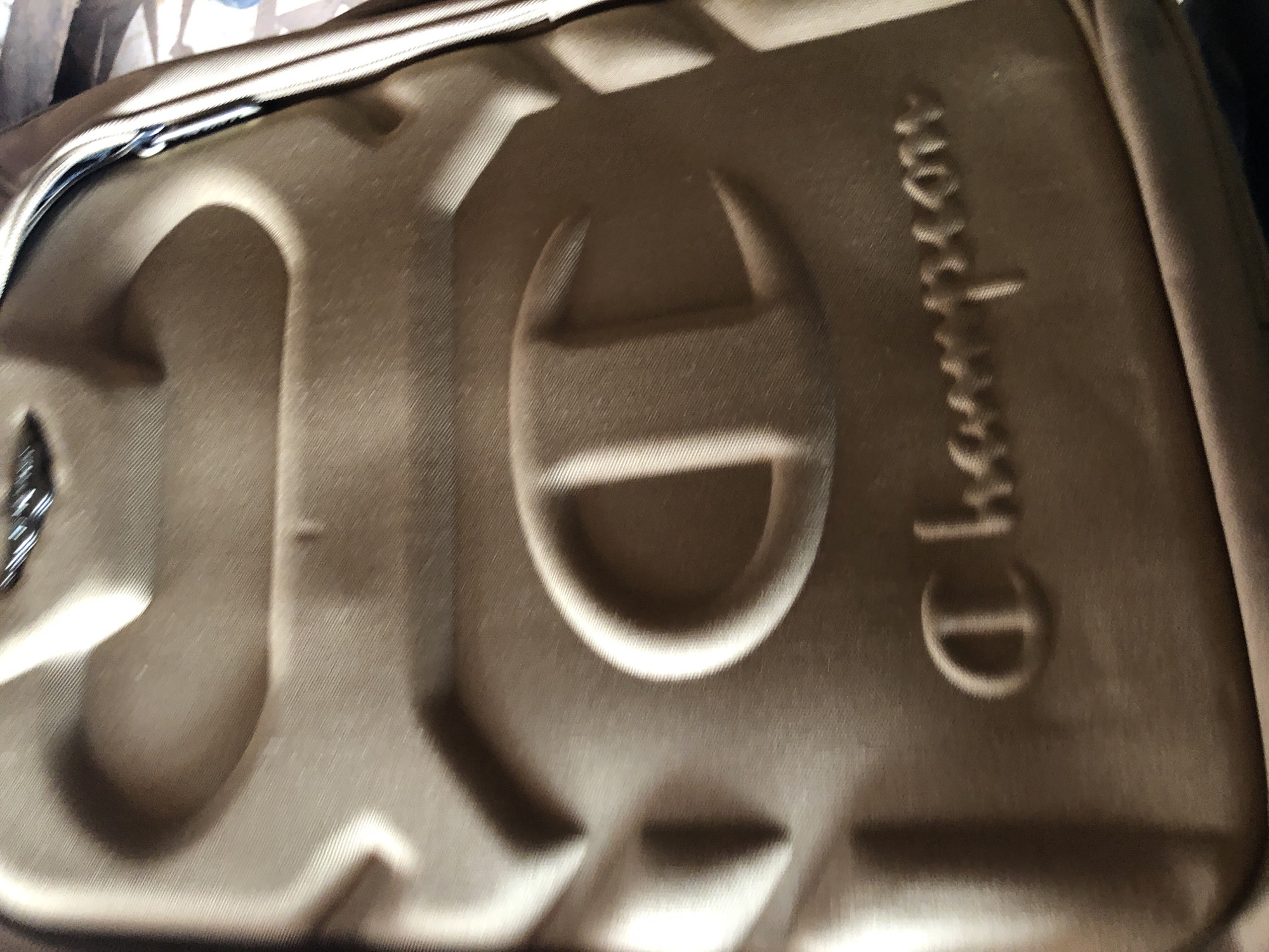 A close-up, rotated shot displays the textured, brownish-khaki or olive drab exterior of a bag or case. The material appears to be fabric, possibly a durable synthetic like nylon or polyester, exhibiting a subtle diagonal weave pattern. Along one edge, a metallic zipper is partially visible.
The surface of the bag features large, raised, embossed elements. Prominently displayed is the iconic "C" logo of the brand Champion, also embossed. Further along the bag, the word "Champion" is embossed vertically, with the letters "hampion" clearly discernible. The embossing creates distinct shadows, highlighting the three-dimensional nature of the branding.
The background is out of focus and indistinct, suggesting an indoor setting with no specific identifying features. There are no people or discernible activities taking place. The time of day and weather cannot be determined from the image.