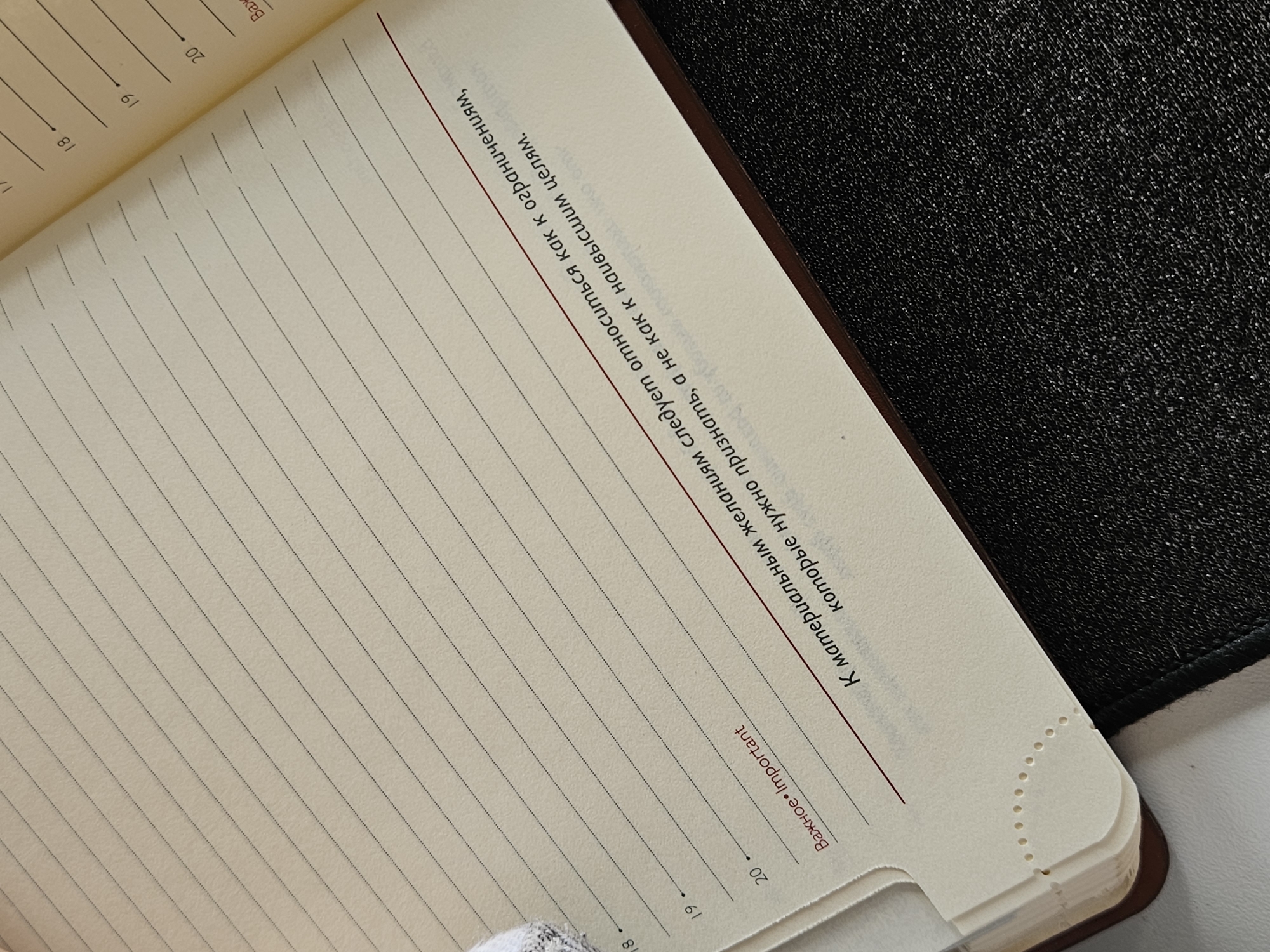 The image shows an open notebook with lined pages and text in Russian. The page is titled "Важное. Important," followed by the text: "К материальным желаниям следует относиться как к ограничениям, которые нужно признать, а не как к наивысшим целям." This translates to "Material desires should be treated as limitations, which must be recognized, and not as the ultimate goals." The notebook is opened to a fresh page with numbered lines, specifically lines 18, 19, and 20 are visible. The notebook's cover is black and textured. Given the Cyrillic alphabet and the name of the city in the instructions, it is assumed the location of this notebook is somewhere in the city of Chaikovsky, Russia.